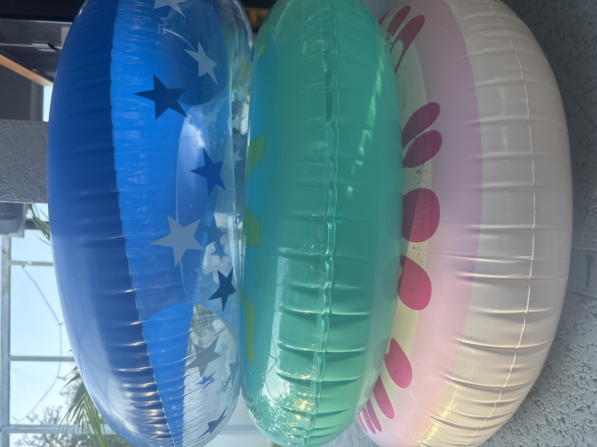 Three inflatable swimming rings are stacked on top of each other outdoors. The top ring is blue with white and dark blue stars. The middle ring is a translucent teal color. The bottom ring is pink and white with pink polka dots. The rings are resting on a gray, textured surface, possibly a patio. In the background, a large window or glass wall is visible, with some greenery and parts of a building structure. The lighting suggests it is daytime. The location of Trinity, United States of America, is not visible in the image.