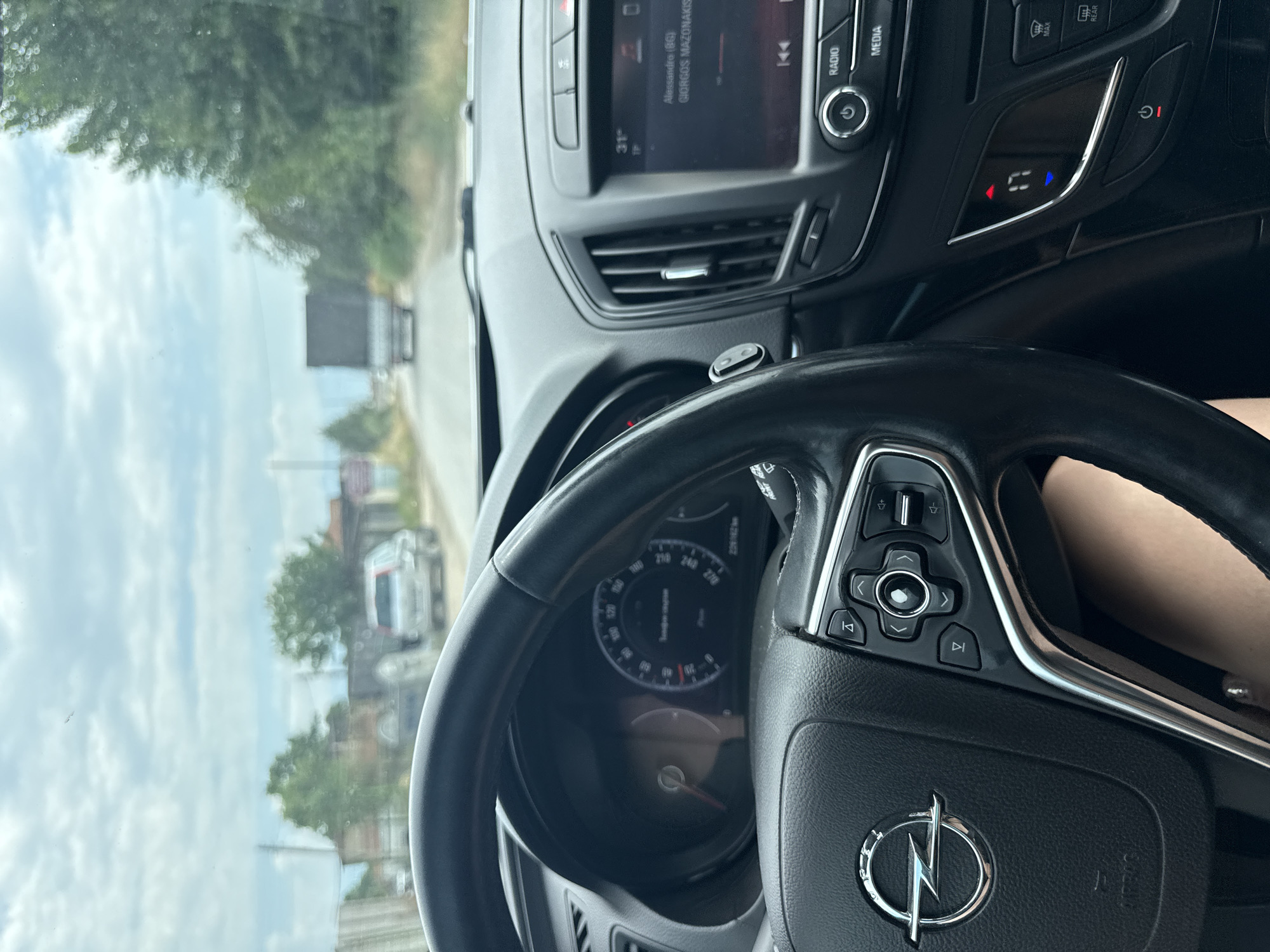 The image is taken from the perspective of someone sitting in the driver's seat of an Opel car. The steering wheel is visible on the left, with the Opel logo clearly displayed. The dashboard features a speedometer and an infotainment screen. The infotainment screen shows the temperature as 31 degrees and is playing music by Alessandro (BG) - GIORGOS MAZONAKIS. The car appears to be on a road in an outdoor environment, with trees and buildings visible in the background. There are other vehicles on the road, including a silver car and a truck. The sky is cloudy. The location is identified as Govedare, Bulgaria.