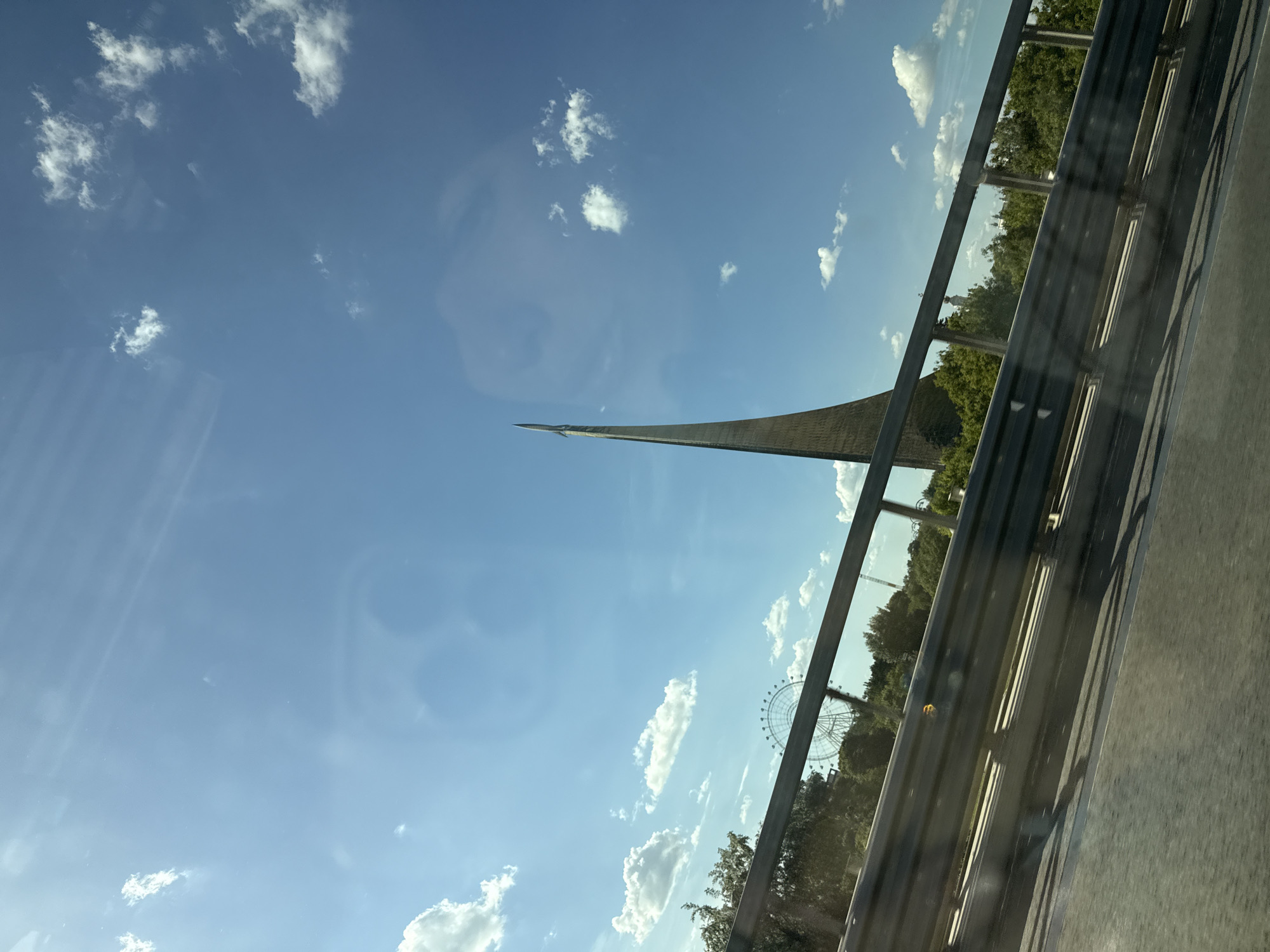 The image, taken from a slightly rotated perspective, depicts an outdoor scene in Moscow, Russia, characterized by prominent landmarks and a clear sky. A dark metal road barrier runs diagonally across the foreground, suggesting the viewpoint is from a vehicle on a road or overpass. Faint reflections, possibly from a window, are visible on the left side of the frame, including a circular outline.
The central feature is the towering Monument to the Conquerors of Space, a sleek, silvery-grey obelisk culminating in a rocket-like structure, pointing skyward. To the right, beyond a stretch of green trees, a large white Ferris wheel, likely the "Sun of Moscow," is partially visible against the horizon. The sky is a bright, clear blue, dotted with scattered white, fluffy clouds.
The scene is bathed in bright daylight, indicating a sunny day, likely mid-day or afternoon. The combination of the distinctive monument and the Ferris wheel strongly identifies the location as the VDNKh area in Moscow. No people are visible, and there is no discernible text within the image. The overall impression is that of a dynamic view of famous city structures from a passing perspective.