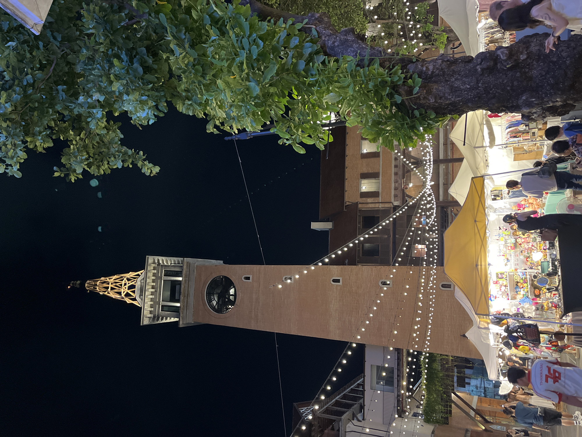 The image depicts a night scene in Chiang Mai City Municipality, Thailand. A tall brick clock tower dominates the left side of the frame, topped with a skeletal metal structure. String lights hang from the buildings and trees, illuminating a busy outdoor market on the right. People are browsing the merchandise displayed under white and yellow awnings. The foreground is partially obscured by tree branches with green leaves.