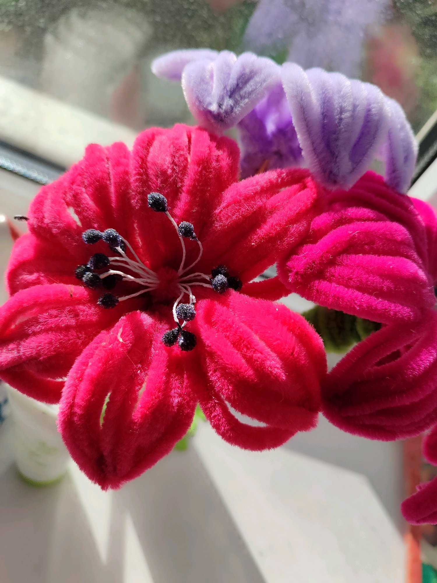 A close-up, top-down view captures two fuzzy, artificial flowers in bright fuchsia and pastel purple. The fuchsia flower is in the foreground and takes up most of the frame. Its petals are wide and curve inwards, with black, textured stamen clustered in the center. The purple flower is partially visible behind the fuchsia one, with its petals gently unfurling. Both flowers appear to be made of pipe cleaners covered in a soft, velvety material. The background is blurred, suggesting a shallow depth of field. Light filters in from the left, creating soft shadows and highlights on the flowers and the white surface they rest on, possibly a windowsill. The overall scene is brightly lit, with a hint of outdoor greenery visible through a window in the top background.