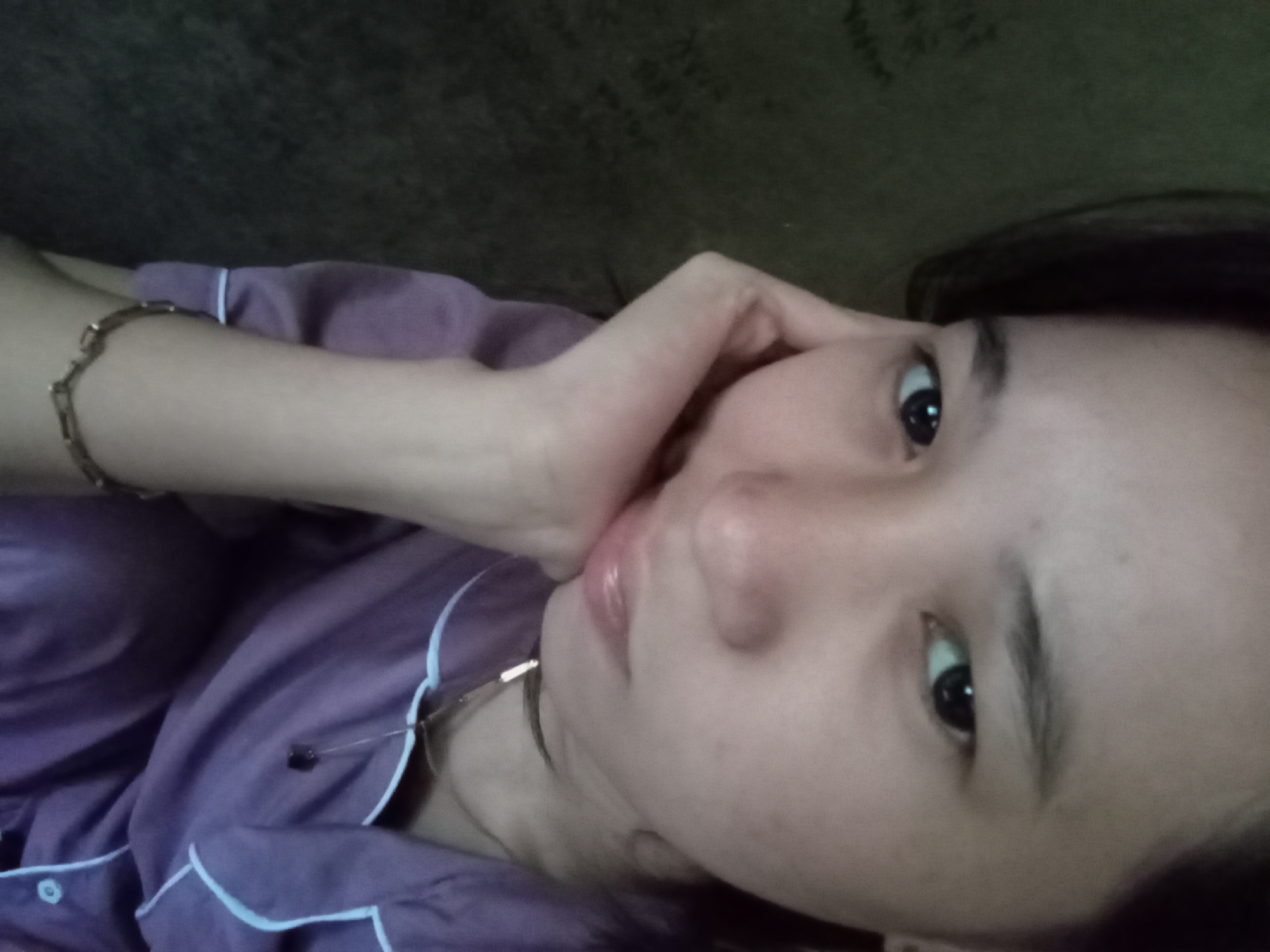 A close-up, frontal, waist-up, indoor, selfie shows a young Asian woman in Rejosari, Indonesia. She is wearing purple pajamas with light blue trim and a simple necklace with a dark pendant. Her left hand supports her chin and cheek, and she is looking directly at the camera. The background is a dark, textured surface, possibly a wall or fabric. The lighting is dim, creating a soft focus effect.