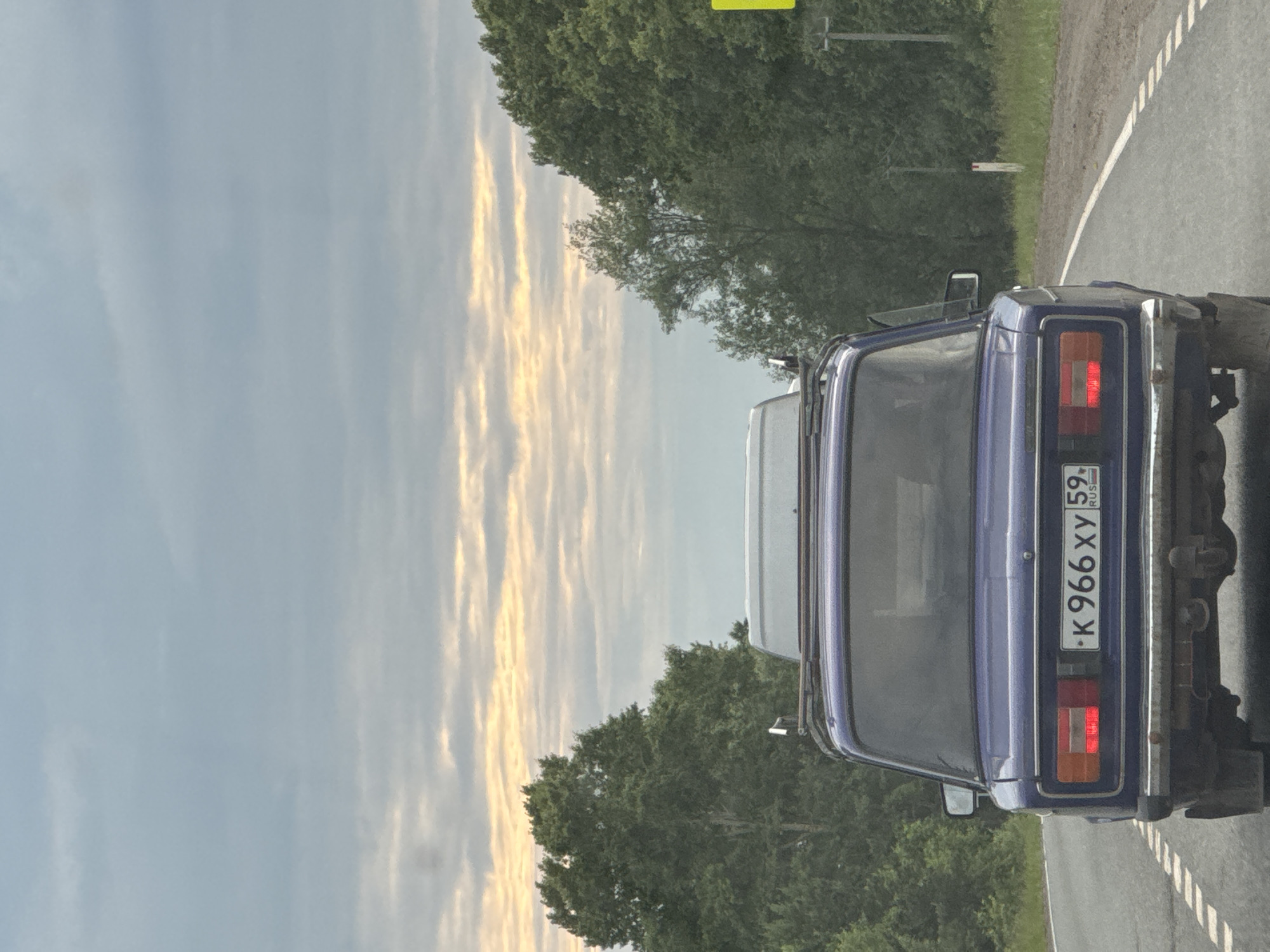 A dark blue sedan, license plate K 966 XY 59 RUS, drives on a paved road with white dashed lines, heading away from the viewer. The car has a roof rack. Trees line the right side of the road, and a yellow sign is visible in the distance. The sky is overcast with streaks of pale yellow light. The scene appears to be in or near the village of Garevaya, Russia.