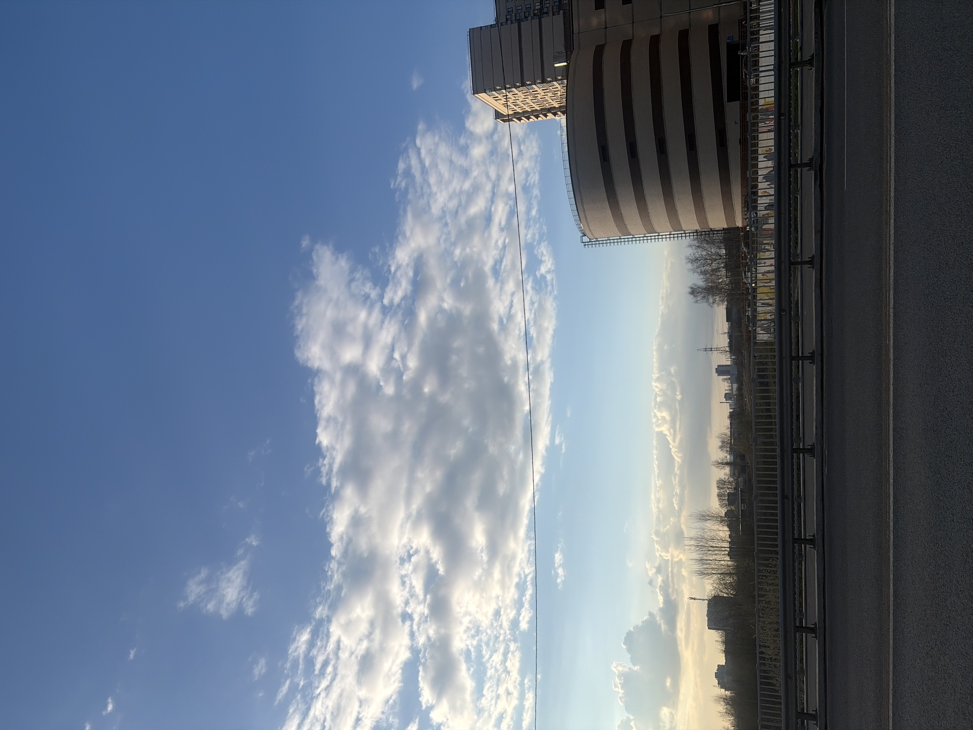 The image shows a view in Saint Petersburg, Russia, featuring a road with a metal guardrail running along its edge. Beyond the railing, there is a strip of trees and bushes with buildings visible in the distance. A modern building with a curved facade and horizontal stripes stands prominently in the scene. The sky takes up a large portion of the image, displaying a mix of blue and clouds. The clouds are dense and white on the left, transitioning to a lighter, wispy appearance towards the right. A thin, dark wire stretches across the sky from the building.