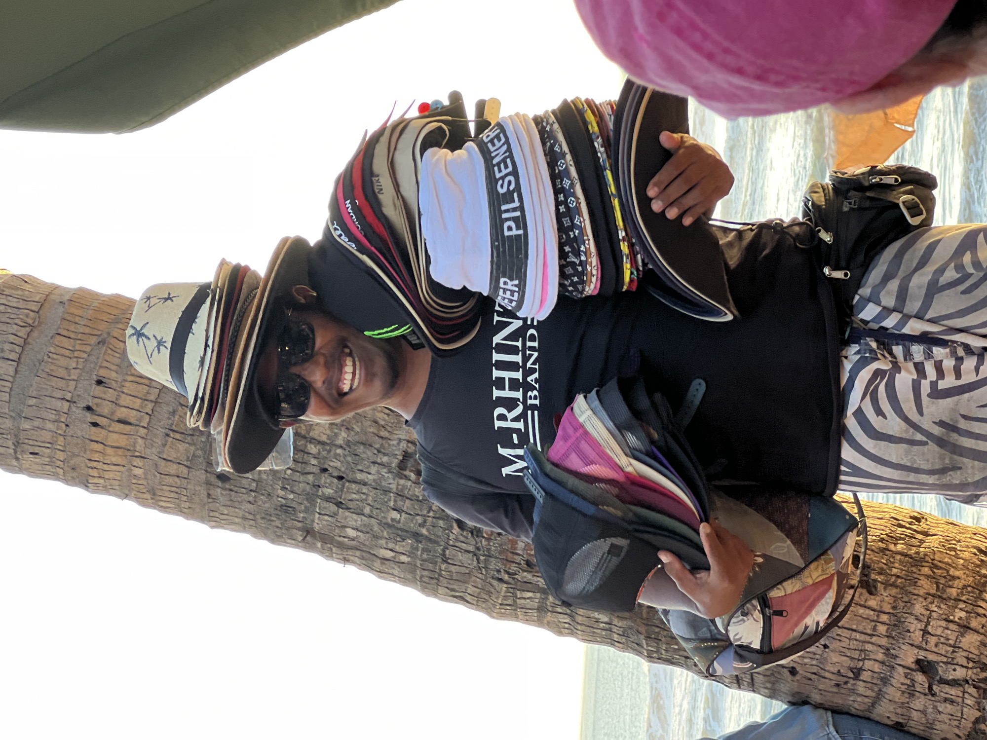 A man stands outdoors near a palm tree and the ocean in Kuta, Indonesia, holding a large stack of hats. He is smiling and wearing sunglasses and a black t-shirt that reads "M-RHIINBEER BAND". He is also wearing patterned pants and a fanny pack around his waist. In his left arm, he holds a bag with various hats inside, and in his right hand, he holds another hat. The sun is setting, casting a warm glow on the scene. A person wearing a pink hat is visible in the foreground.