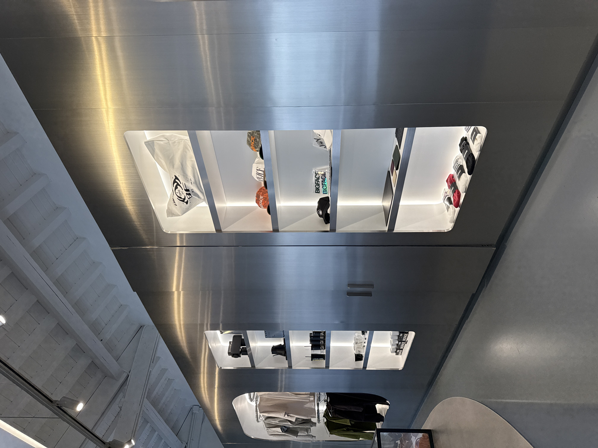 The image shows a mirrored ceiling with display cases embedded within it. The display cases contain items such as hats, t-shirts, and other clothing items. The ceiling is metallic and reflects the interior of what appears to be a store. The lighting is bright, highlighting the items on display. The background contains elements of the store's interior design, including a white ceiling and a concrete floor. It is likely a retail space located in a modern or upscale area. Without additional evidence it cannot be concluded that this is Miami, Florida.