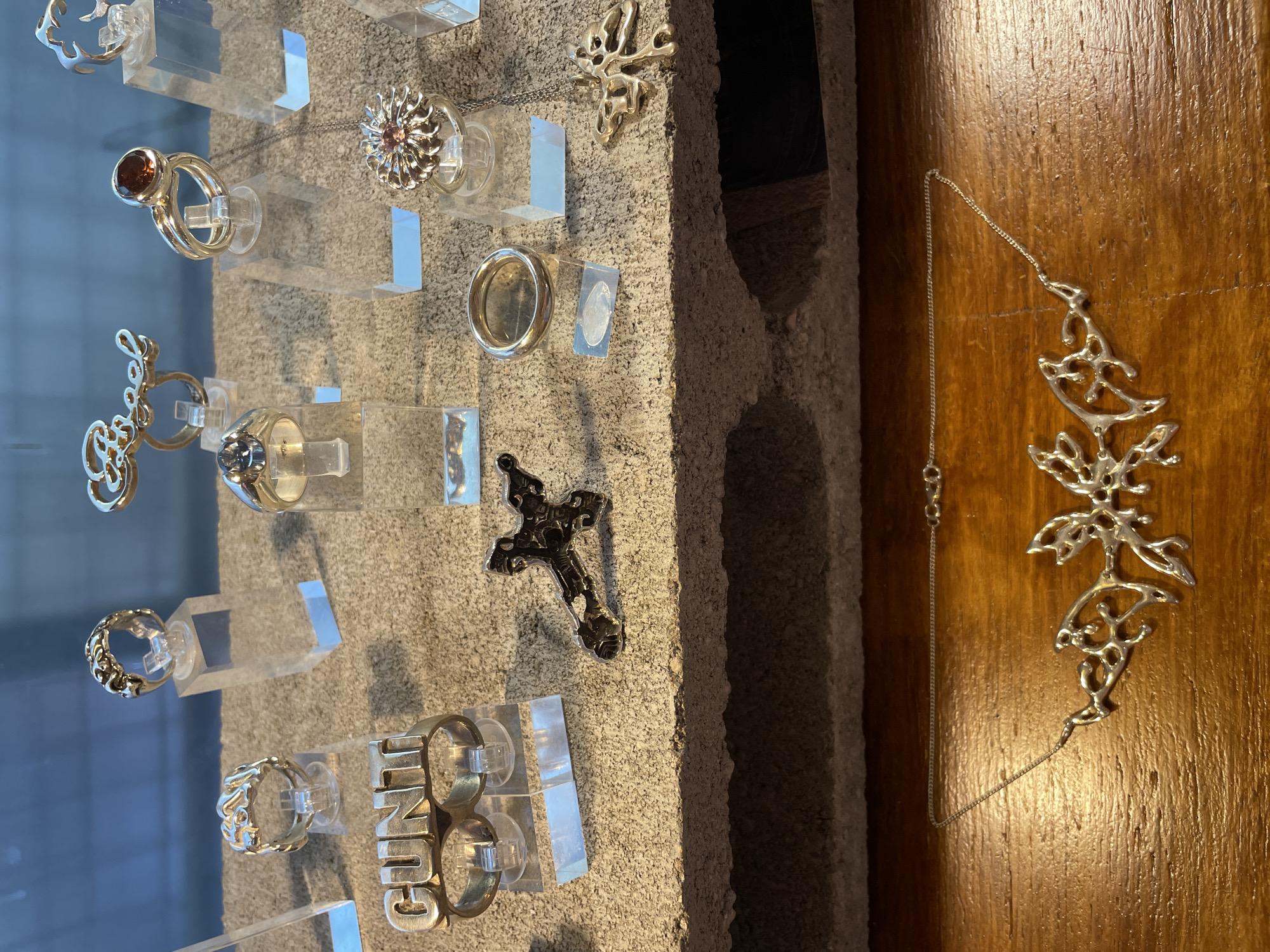 This media file captures a detailed display of handcrafted jewelry, likely in a shop or gallery setting in Buenos Aires, Argentina. The primary subjects are various silver-toned rings, pendants, and a necklace, exhibited against contrasting textures.
The majority of the jewelry is showcased on clear, transparent acrylic risers, which are mounted on a light gray, rough, concrete-like wall. The rings display a variety of designs: one features a prominent reddish-brown oval stone; another has a detailed 'sunburst' or floral pattern with a small central gemstone; a third exhibits an intricate, organic, or abstract carved pattern; and a distinctive ring is shaped to spell "GUNGUN" across two finger holes. Other rings include a simple, smooth band and one with a subtle blue or gray stone.
Among the rings and also affixed to the textured wall are several pendants. One is an elaborate, organic, and abstract shape, while another is a traditional, ornate Christian cross. A small, plain, circular pendant is also visible.
To the right side of the image, a long, delicate silver chain necklace is draped against a warm, richly toned wooden surface, which appears to be a wall panel or door. The necklace features a large, open-work pendant with an intricate, organic, and abstract design, complementing the style seen in some of the other pieces.
The scene suggests an artistic display, emphasizing the unique craftsmanship of the jewelry. The lighting is internal, highlighting the metallic sheen and details of the pieces. No people are visible, and the activity is solely the presentation of the items. The only visible text is the word "GUNGUN" on one of the rings.