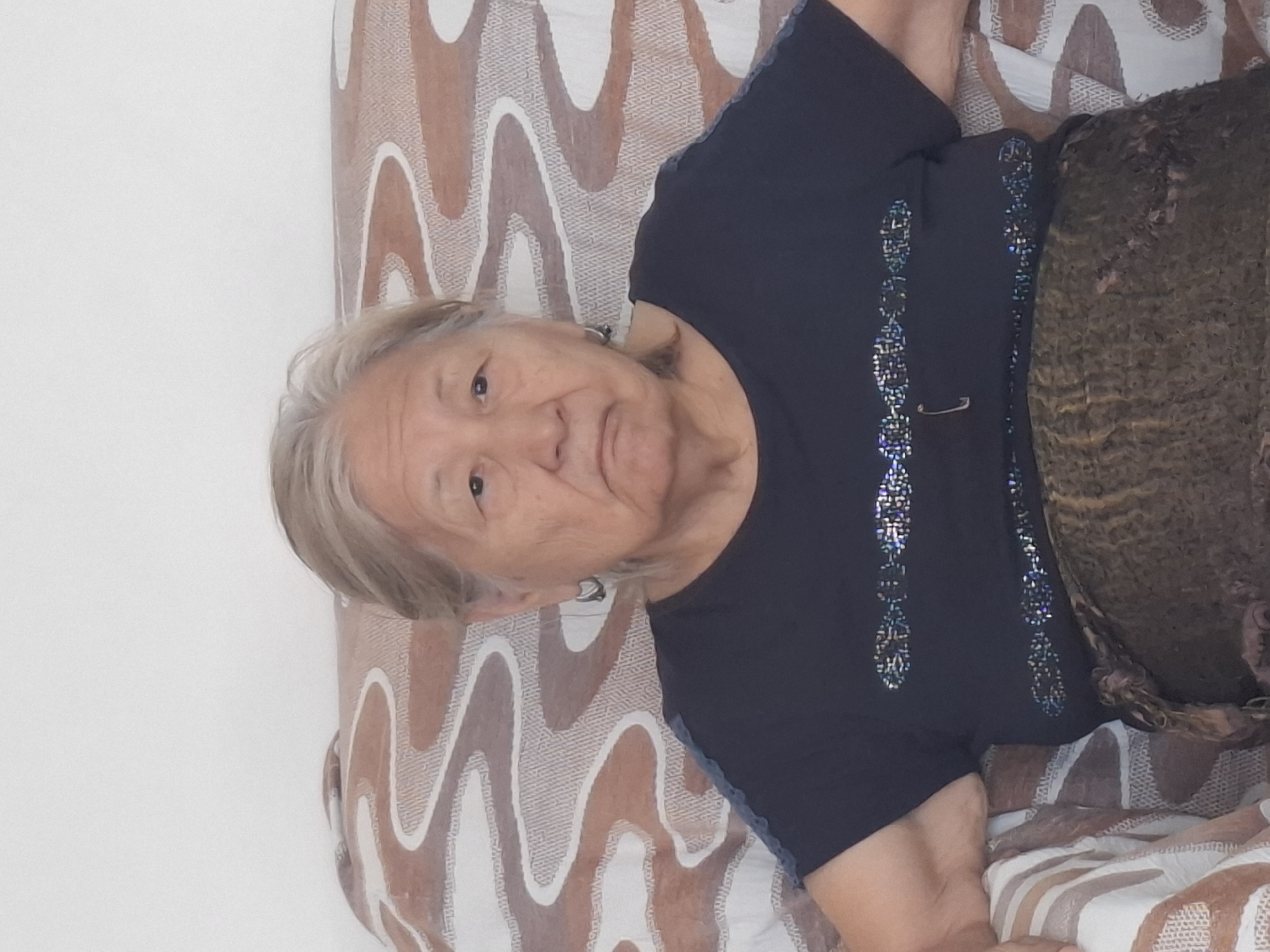 A waist-up, medium shot shows an elderly East Asian woman with graying hair, wearing a dark blue t-shirt with a patterned embellishment and a brown patterned skirt. She is seated on a sofa with a brown and white wavy pattern on the upholstery. The background is a plain white wall. The lighting is soft and even, suggesting an indoor setting. There are no visible indicators of the location within Almaty, Kazakhstan. The woman's expression is neutral.