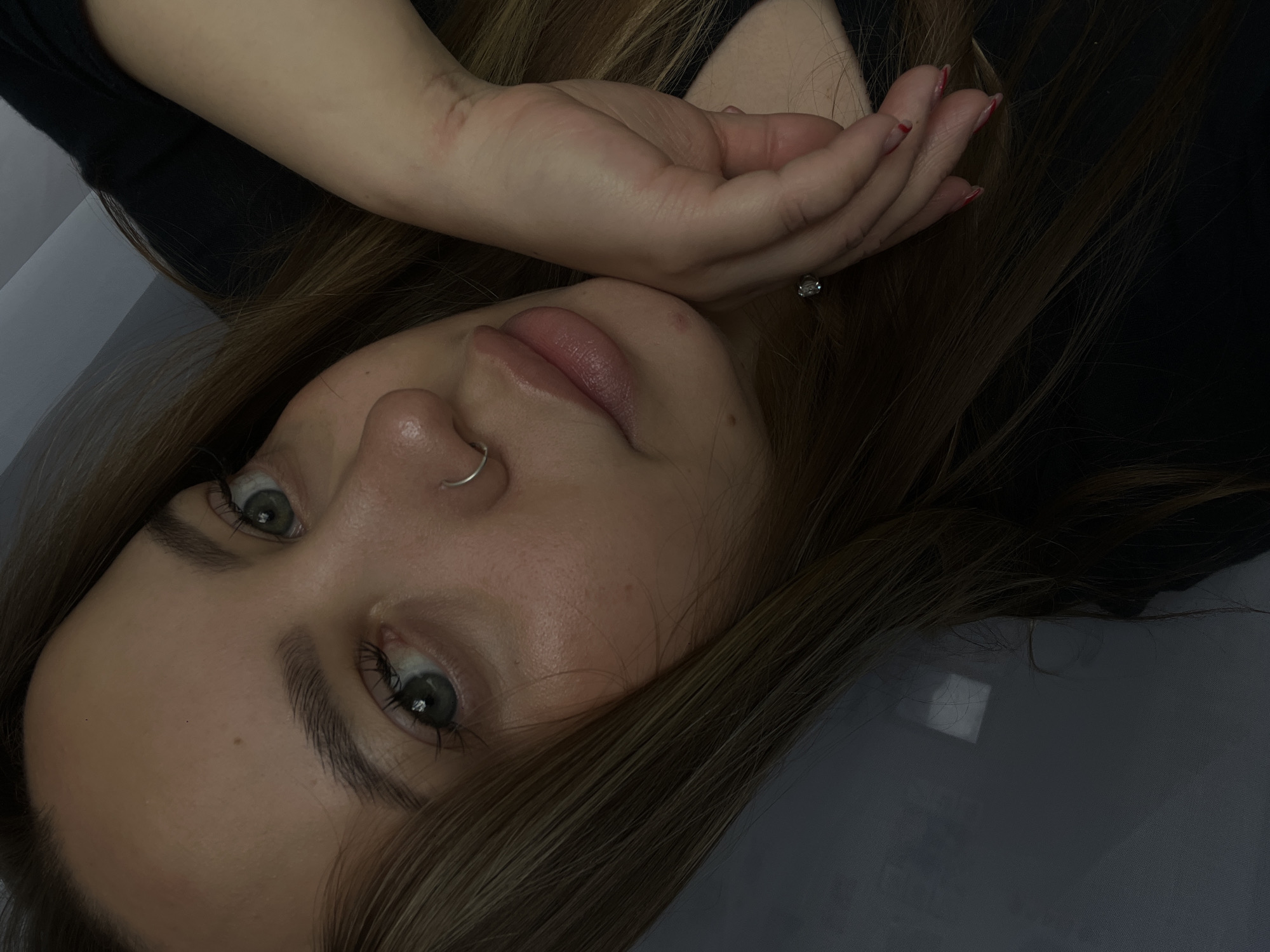 A close-up, downward-angled shot depicts a young woman with light brown hair, fair skin, and striking green eyes. She is lying down, with the back of her right hand gently resting on her forehead near her hairline. She has a nose ring and what appears to be a diamond ring on her right ring finger. Her lips are full and slightly parted. She is wearing a dark-colored top. The background is a muted gray, possibly a sheet or pillow. There is no evidence to determine the location of the city of Novopokrovskaya, Russia in the image.