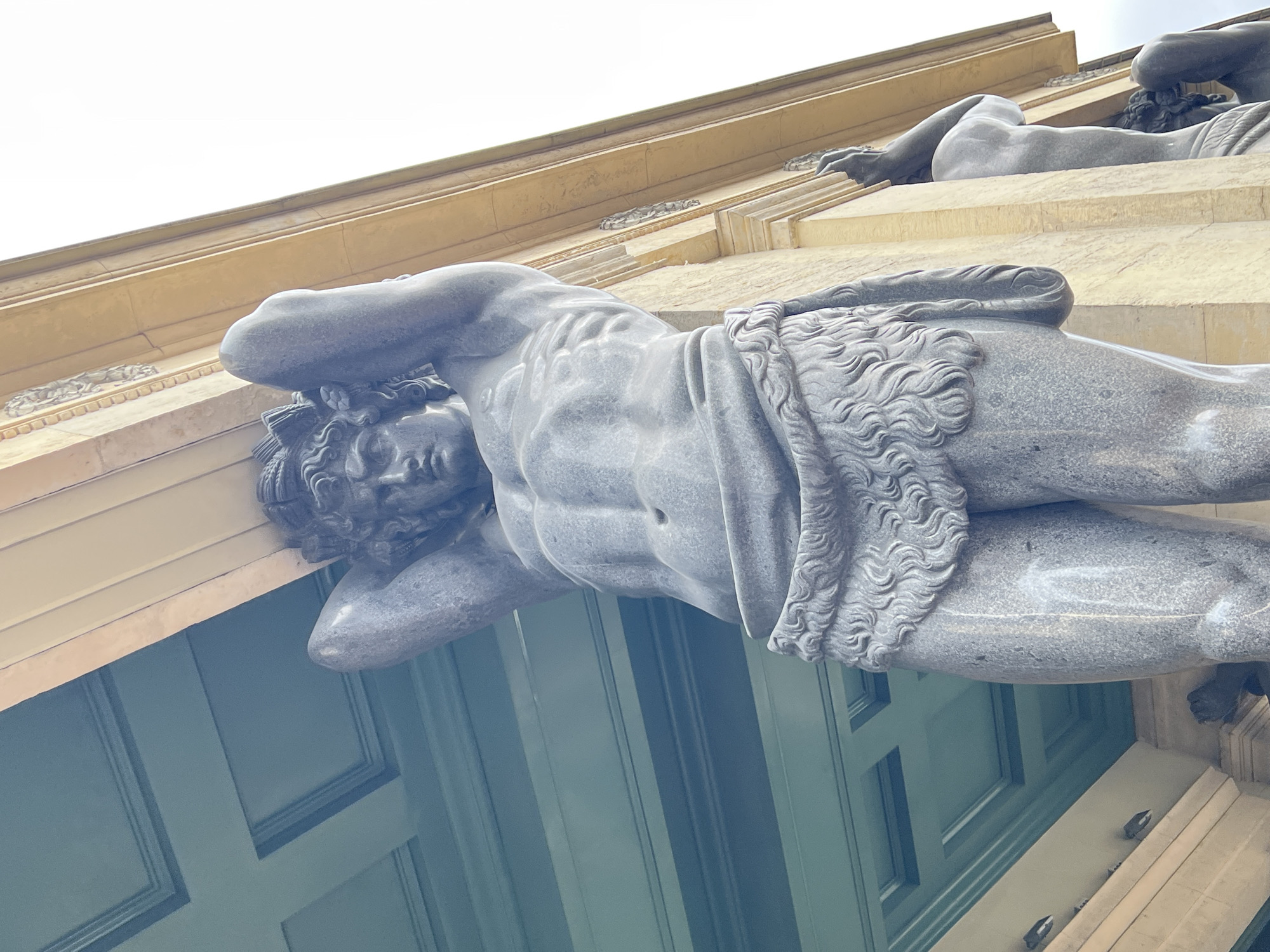 The image shows a low-angle shot of the Atlantes figures supporting the building of the New Hermitage Museum in Saint Petersburg, Russia. The Atlantes are muscular male figures made of granite, depicted with their arms raised, holding up the entablature of the building. The foreground figure is partially visible, showcasing its detailed musculature and a kilt-like garment made of fur. The background reveals the architectural elements of the New Hermitage, including the yellow facade and a teal-colored ceiling with recessed panels. Another Atlantes figure is visible in the background to the right. The sky is bright and overcast.