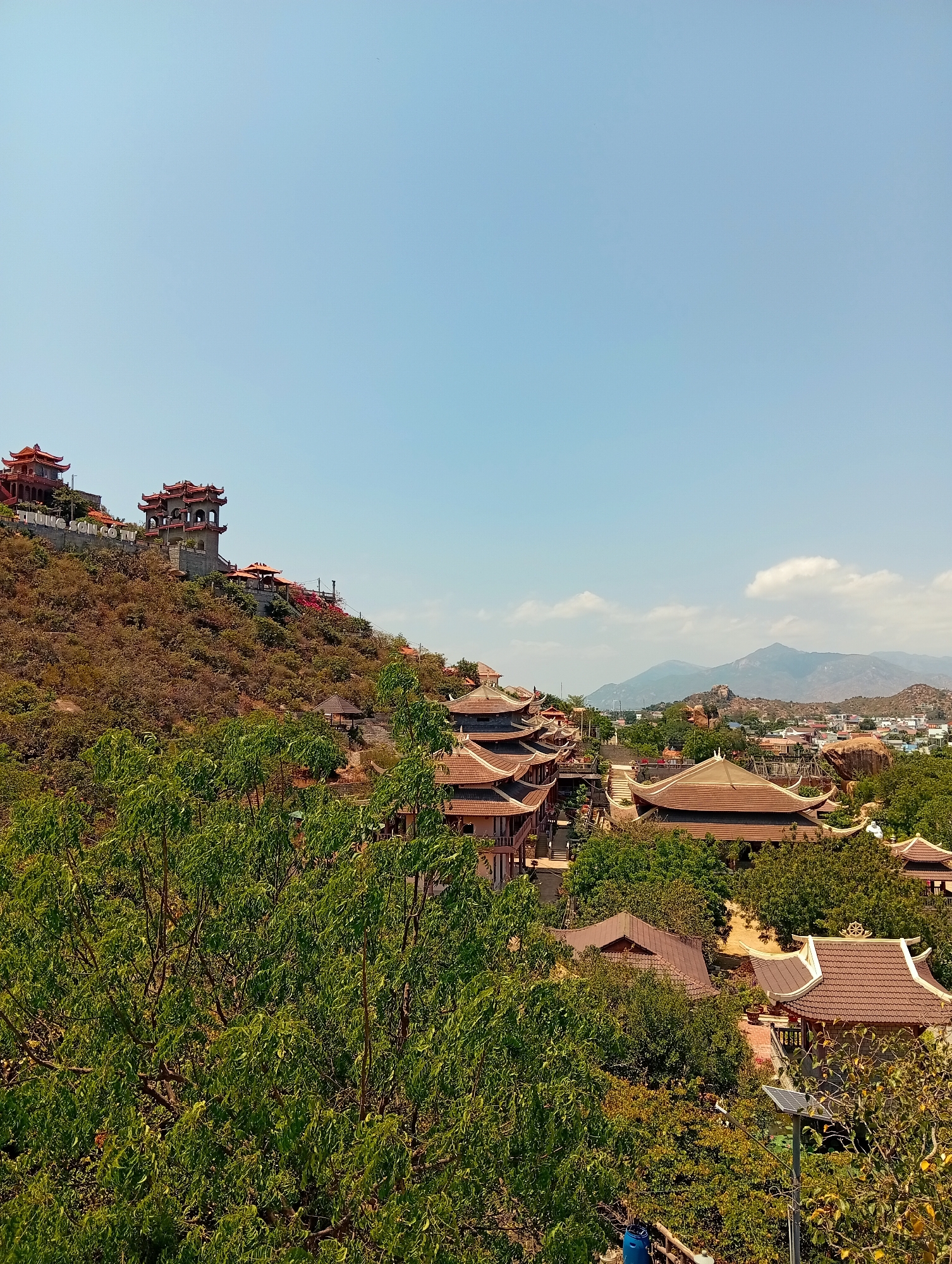 The image depicts a panoramic view of Khánh Hải, Vietnam, showcasing its urban landscape intertwined with natural terrain. In the foreground, lush green trees fill the lower portion of the frame, partially obscuring buildings. Several traditional Vietnamese-style buildings with red-tiled roofs and intricate architectural details are visible. Further up the landscape, a hill rises, dotted with more of the same style of buildings. At the top of the hill, two red-roofed pagodas stand prominently against the clear blue sky. In the background, distant mountains rise against the horizon. The city itself is nestled amidst the trees and hills, with glimpses of rooftops and buildings suggesting a settled urban presence. A solar panel and lamp post stand within the foreground vegetation.
