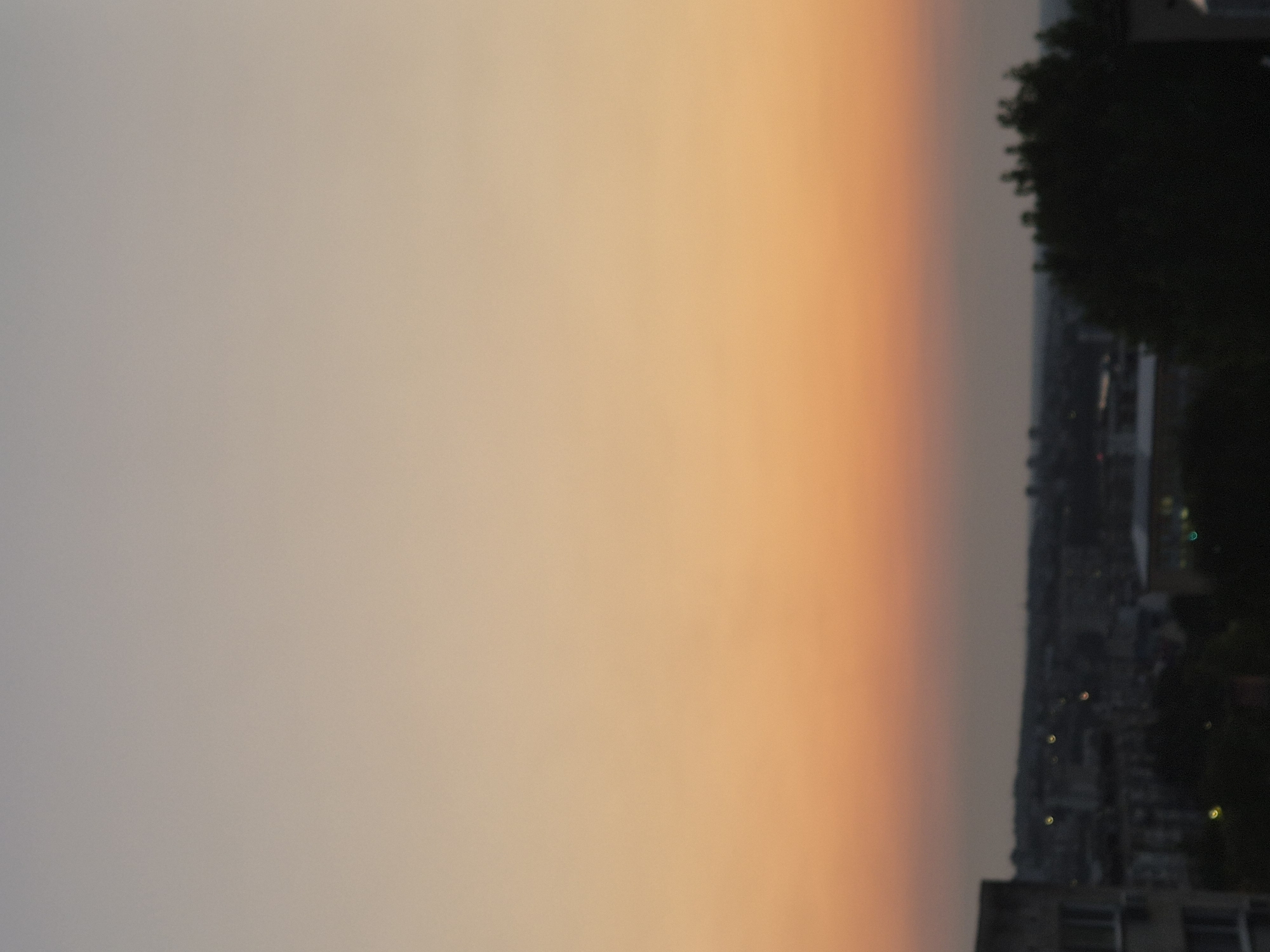 An aerial, eye-level shot captures a cityscape at dusk. The sky transitions from a light gray at the top to a warm orange and then a muted pink near the horizon. Silhouetted against the dusky sky are buildings and trees that form the cityscape. Some of the buildings are tall and rectangular, while others appear shorter and more spread out. There are also several trees, their dark foliage obscuring details of the city below. Scattered throughout the cityscape are small, blurred lights, suggesting that some of the buildings are illuminated. The overall impression is one of a quiet evening in an urban environment.
