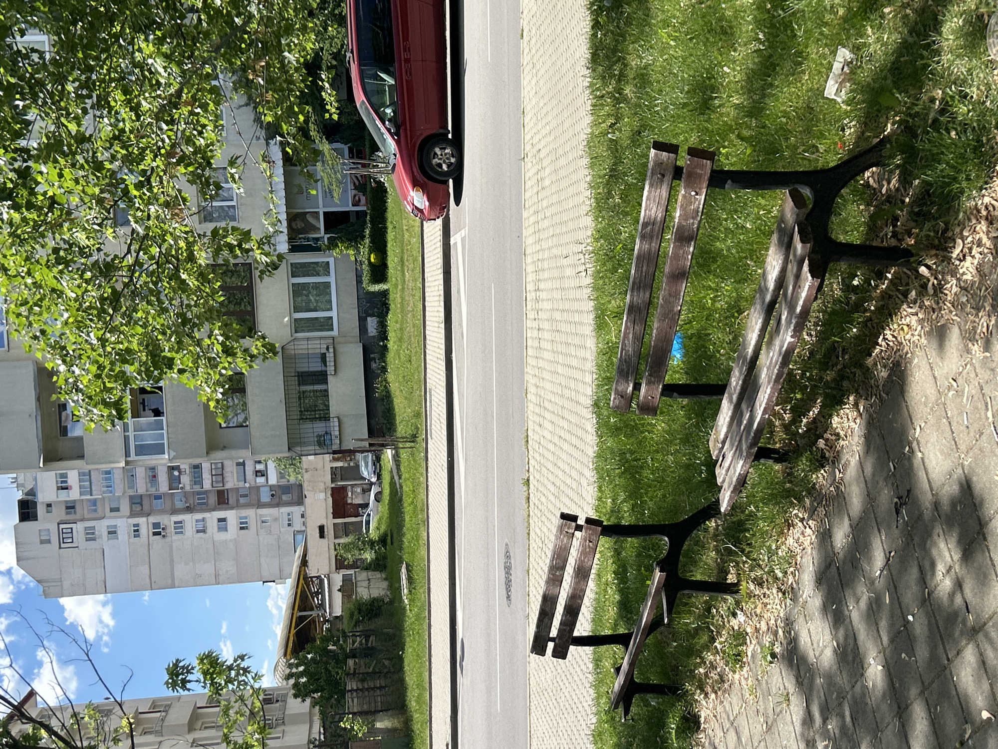 The scene is an urban environment. A street runs between a tall building on one side and a grassy area with two benches on the other. A red car is parked alongside the road. The benches are wooden with dark supports and are situated on a paved area next to the grass.