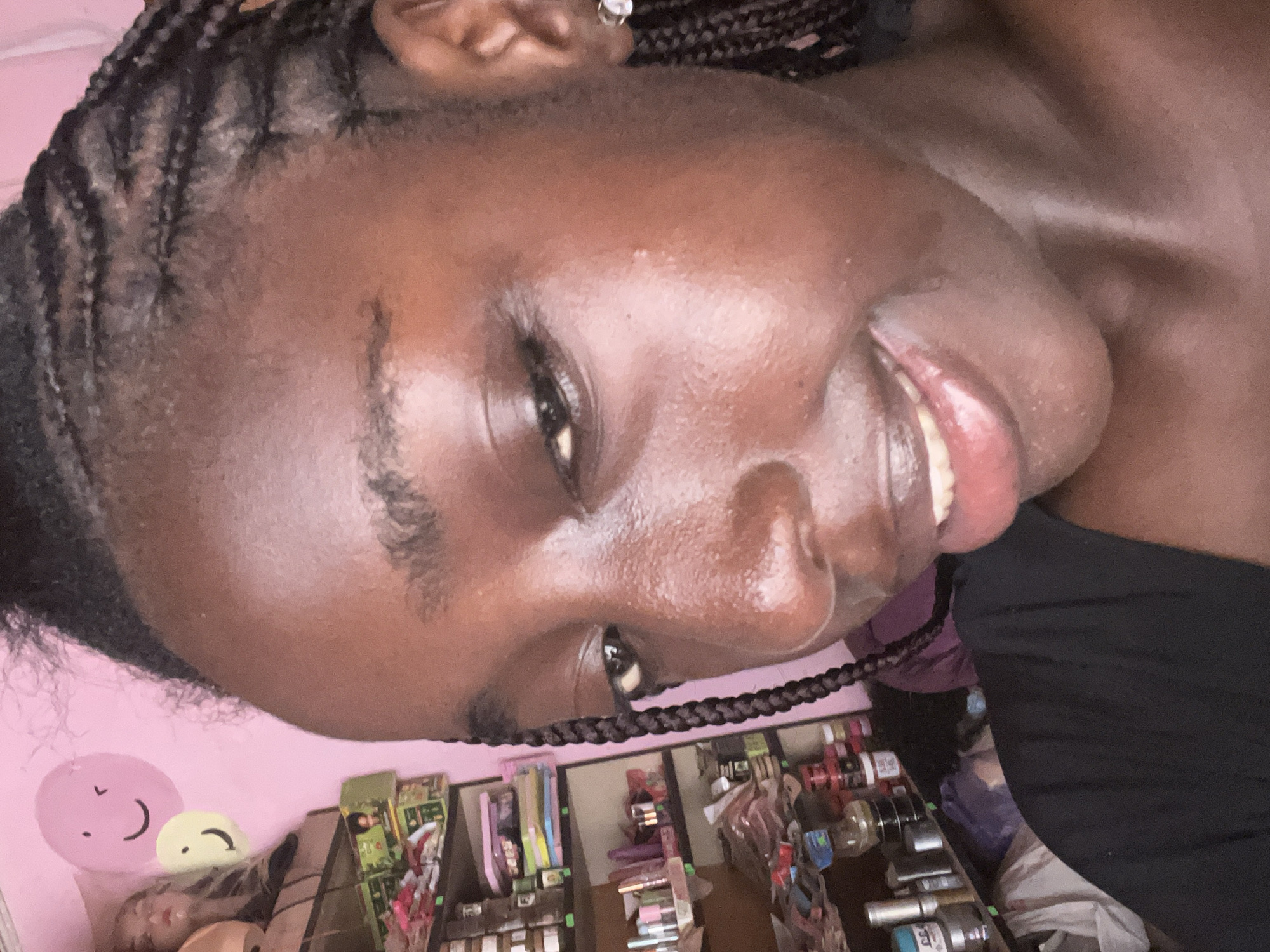 The image is a close-up selfie of a person with dark skin and braided hair. The person is smiling slightly, revealing their teeth. The background is blurry but appears to show a room with a pink wall, a shelf or table filled with various objects, possibly cosmetics or beauty supplies, and some decorative items on the wall. It is impossible to definitively determine the location as Abuja, Nigeria, based on the image alone. There is no identifying information to support that location.