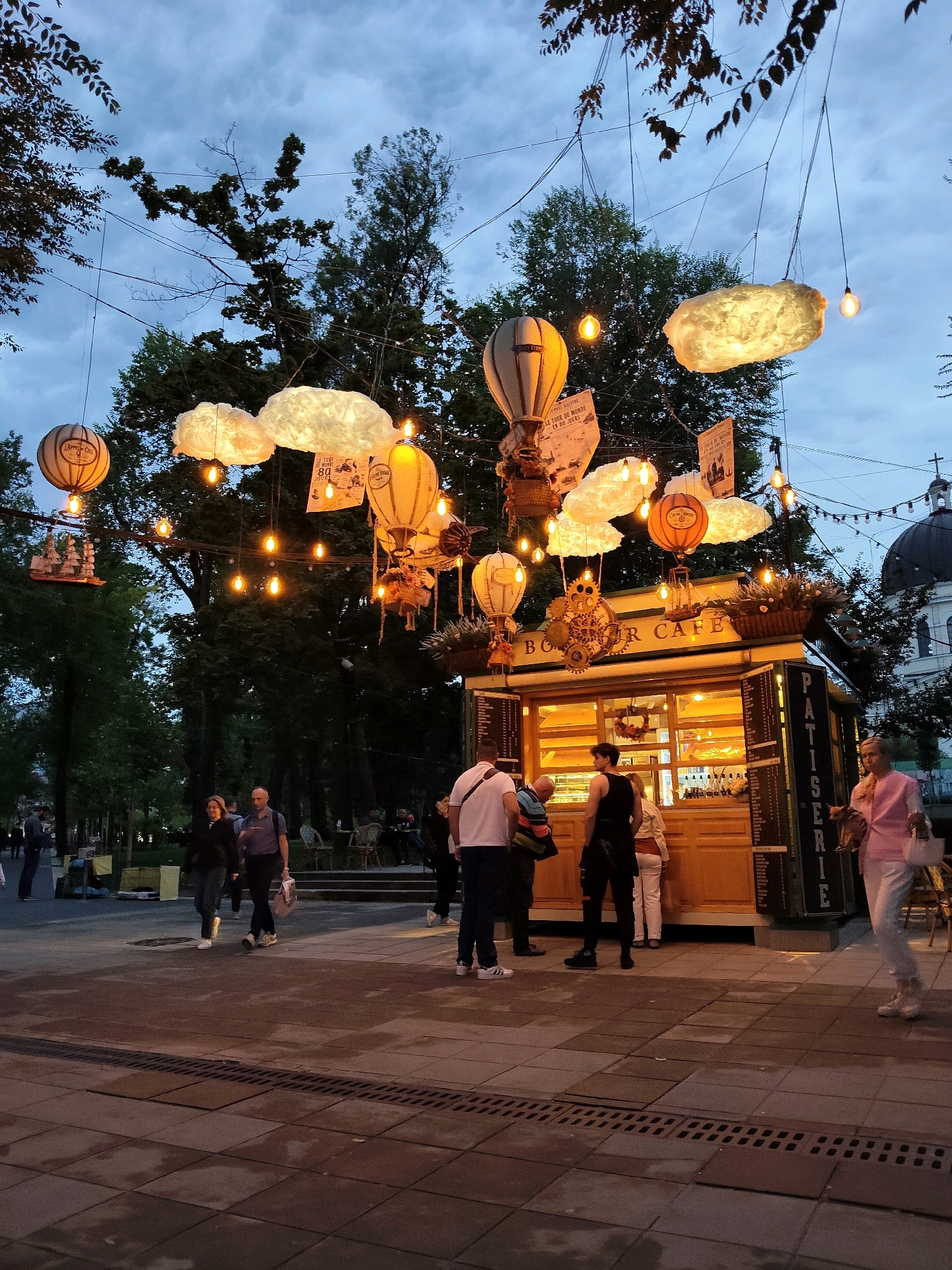 The image depicts an outdoor scene at dusk, likely in a public park or pedestrian area in Chișinău, Moldova. The focal point is a charming, illuminated "BOUCHER CAFE" kiosk, designed with a rustic wooden facade and large display windows showcasing baked goods. The cafe is adorned with whimsical decorations, including hot air balloon-shaped lanterns, fluffy cloud-like lamps, and various hanging ornaments, creating a festive atmosphere. String lights are draped across the trees and overhead, casting a warm glow.
Several people are visible interacting with the cafe and walking by. In the foreground, a group of individuals stands near the counter, some appearing to order or receive items. Further back, a couple walks together on a paved walkway. In the background, more people are seated at tables, suggesting a relaxed social gathering. The environment includes mature trees, a paved walkway with decorative patterns, and a glimpse of a building with a domed roof in the distance, which could be a church or other historical structure. The sky is overcast with twilight hues.