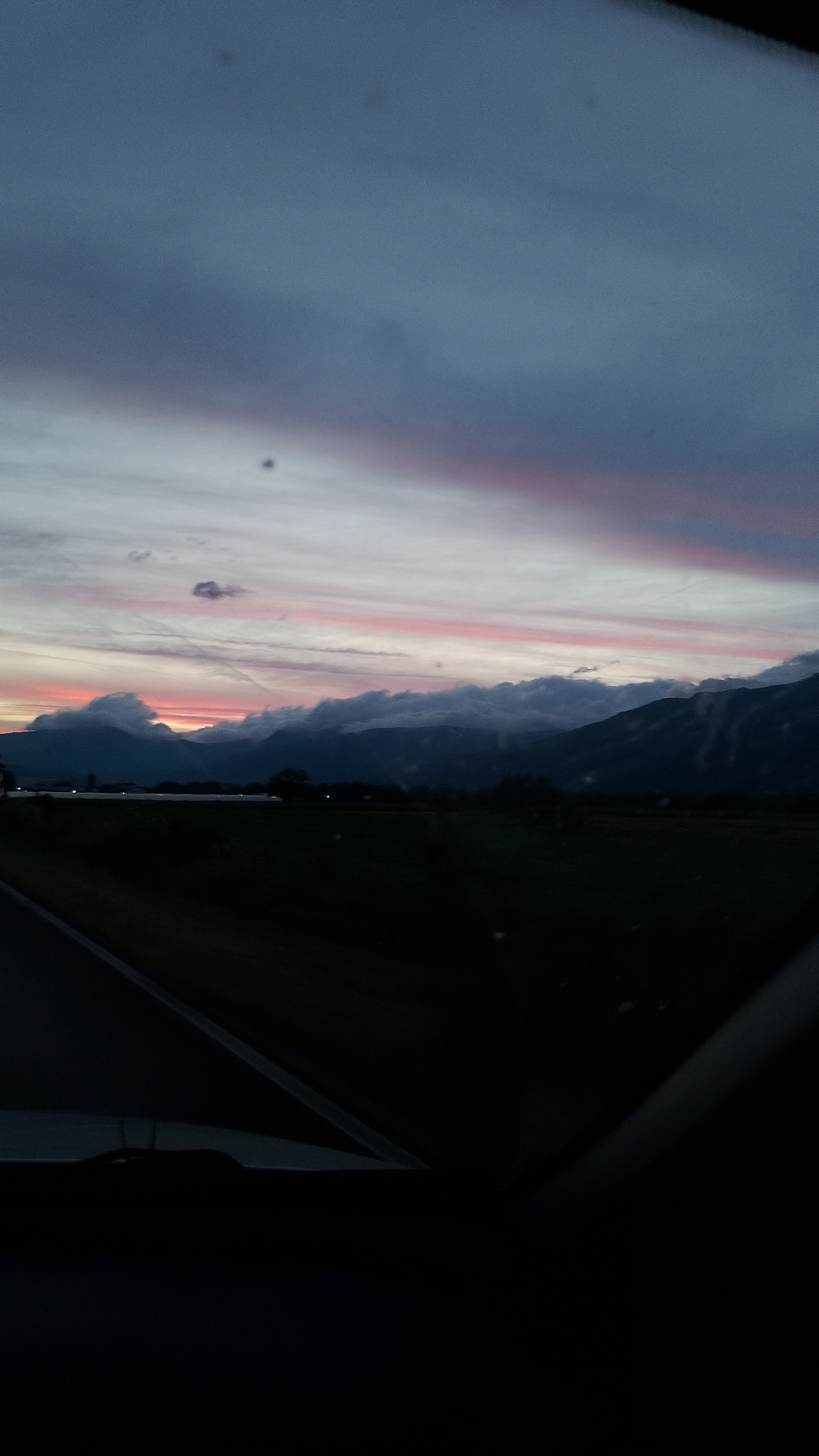 The image captures a twilight scene with a dramatic sky over a landscape. In the foreground, a dark road curves to the left, with a hint of a car's interior visible along the bottom edge. The middle ground is dominated by a dark, silhouetted field of grass. Beyond the field, a range of mountains rises, partially obscured by heavy, dark clouds that are illuminated from below by the fading sunlight. The sky above the mountains displays streaks of pink and orange, transitioning into a deep blue at the top of the frame. A few small, dark spots are visible on the sky. The overall impression is one of a serene, natural landscape at dusk, with the mountains of Gabarevo, Bulgaria, providing a majestic backdrop.