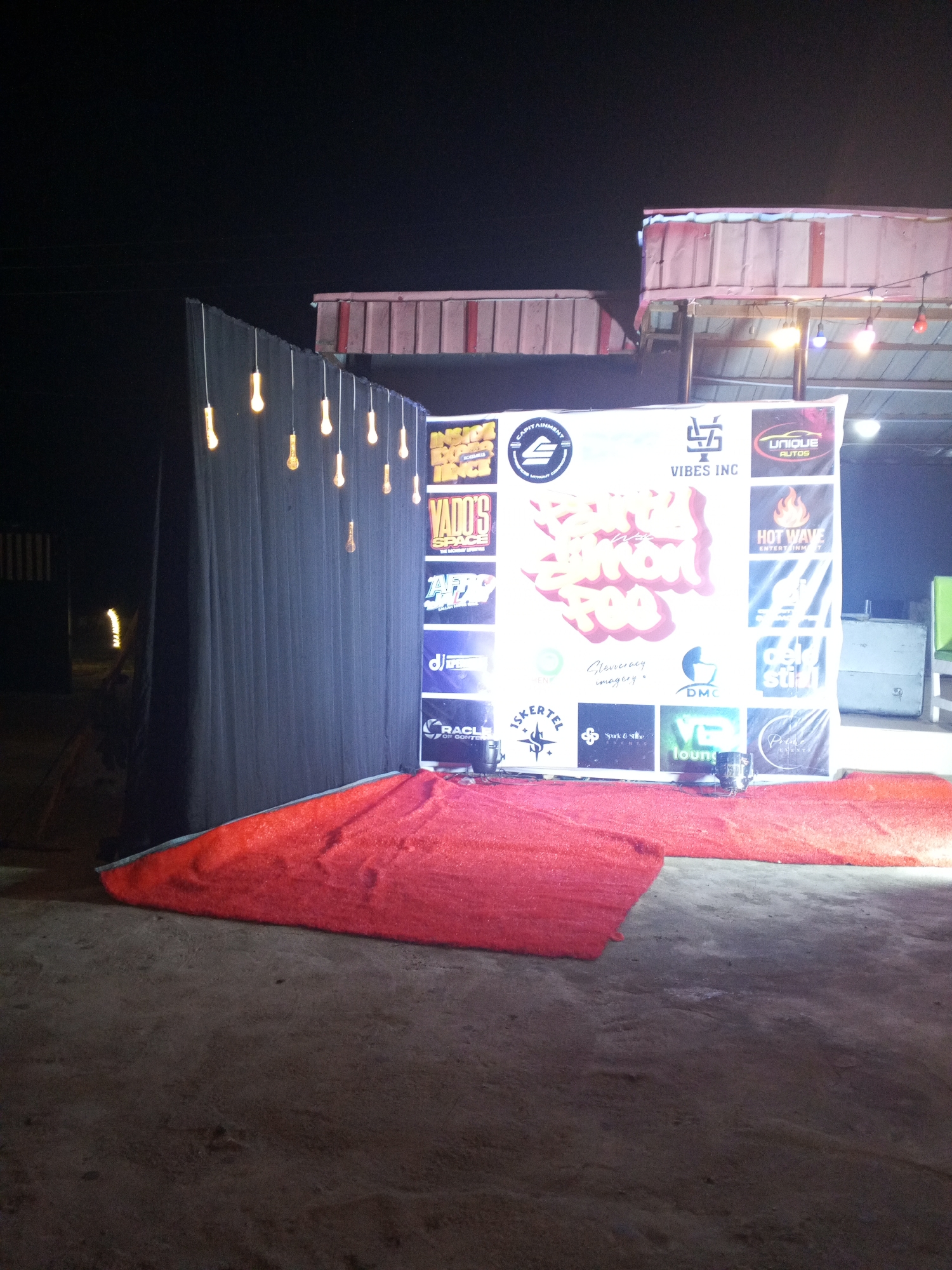The image shows a makeshift event stage. A black backdrop with hanging light bulbs sits to the left, while a sponsor banner is on the right. A red carpet covers the area in front of the backdrop and banner. The location is presumed to be Lokoja, Nigeria, based on the logos visible.
