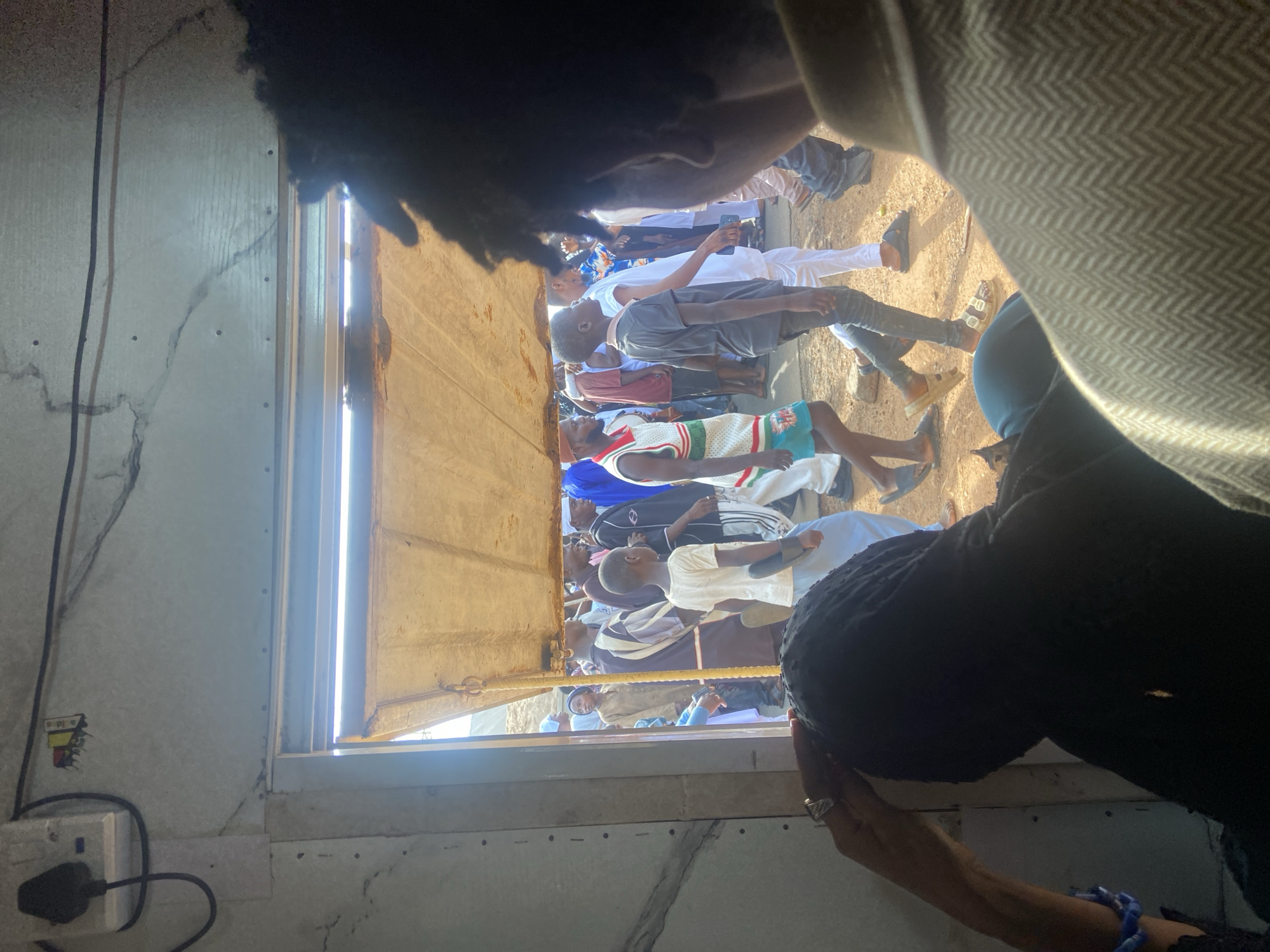 The image depicts a view from inside a building, looking out a window or opening, at a group of people gathered outside. The location is Okengwe - Obehira, Nigeria.
Several individuals are visible outside, many standing or sitting on the ground. Some are wearing casual clothing, including jerseys and T-shirts. Some are barefoot.
The foreground contains elements of the building's interior, including a power outlet, wall, and framing around the opening. There is also a dark shape obscuring part of the view, possibly hair or clothing. An arm and hand are visible in the bottom right corner.