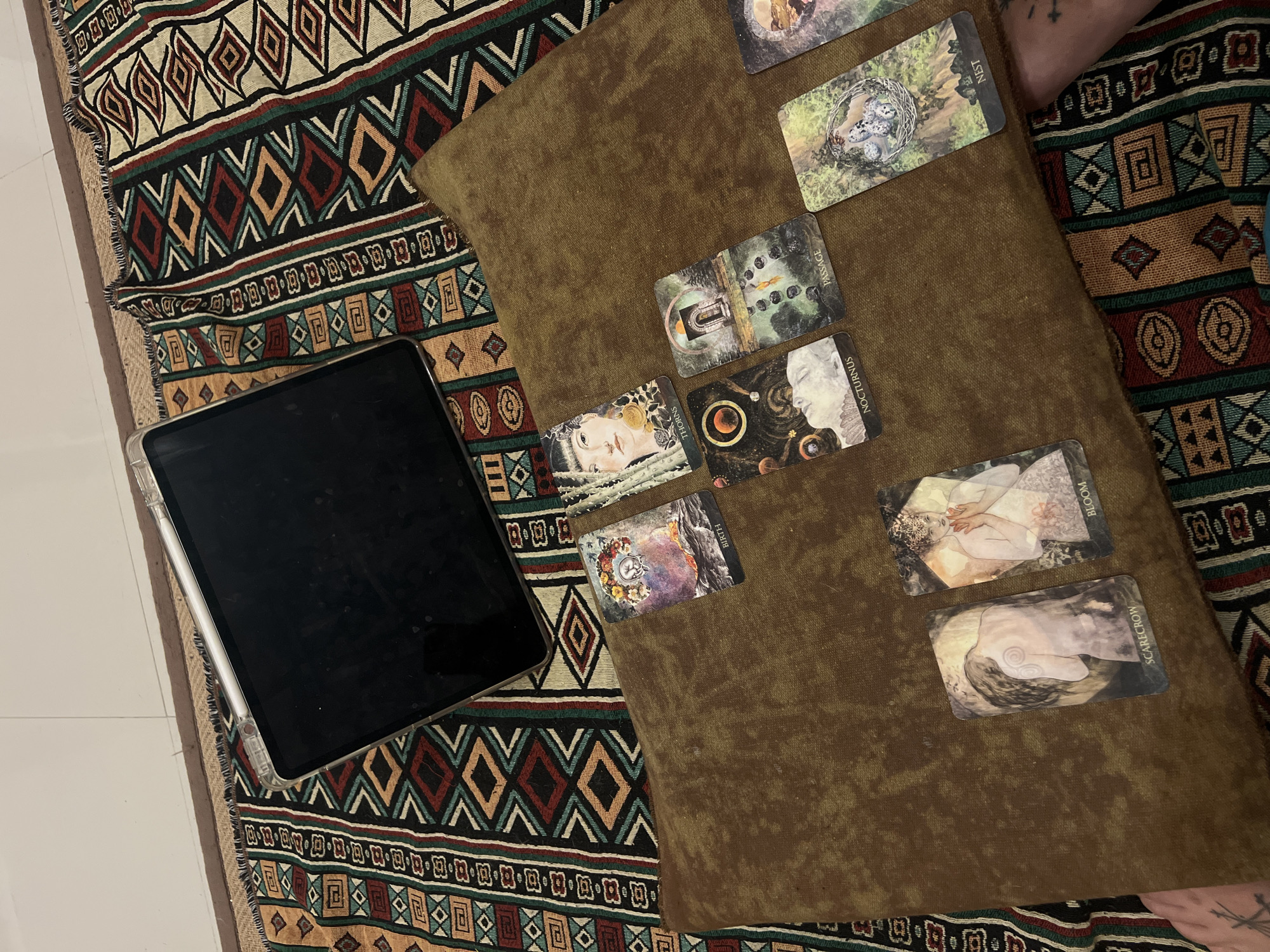 The image shows a flat lay of a tablet and several tarot cards arranged on a patterned rug and a brown cloth. The tablet is positioned on the left side of the image, covering a portion of the rug. The tarot cards are scattered across the brown cloth and the rug. Some of the cards have visible text labels, including "NEST," "PASSAGE," "THORNS," "NOCTURNUS," "BIRTH," "BLOOM," and "SCARECROW." The tarot cards feature artistic illustrations, some with figures and nature imagery. The background rug has a geometric pattern with diamond shapes in various colors. The setting appears to be indoors, possibly a living space, with tiled flooring visible to the left of the rug. There are no people directly visible in the frame, but a faint tattoo is visible on the upper right corner of the image. The overall impression is a mystical or introspective scene, possibly related to a tarot reading. The image is well-lit, suggesting daylight, although no specific time of day can be definitively determined. No weather cues are present. The location context of Da Nang, Vietnam, does not provide any specific visual cues within the image itself.