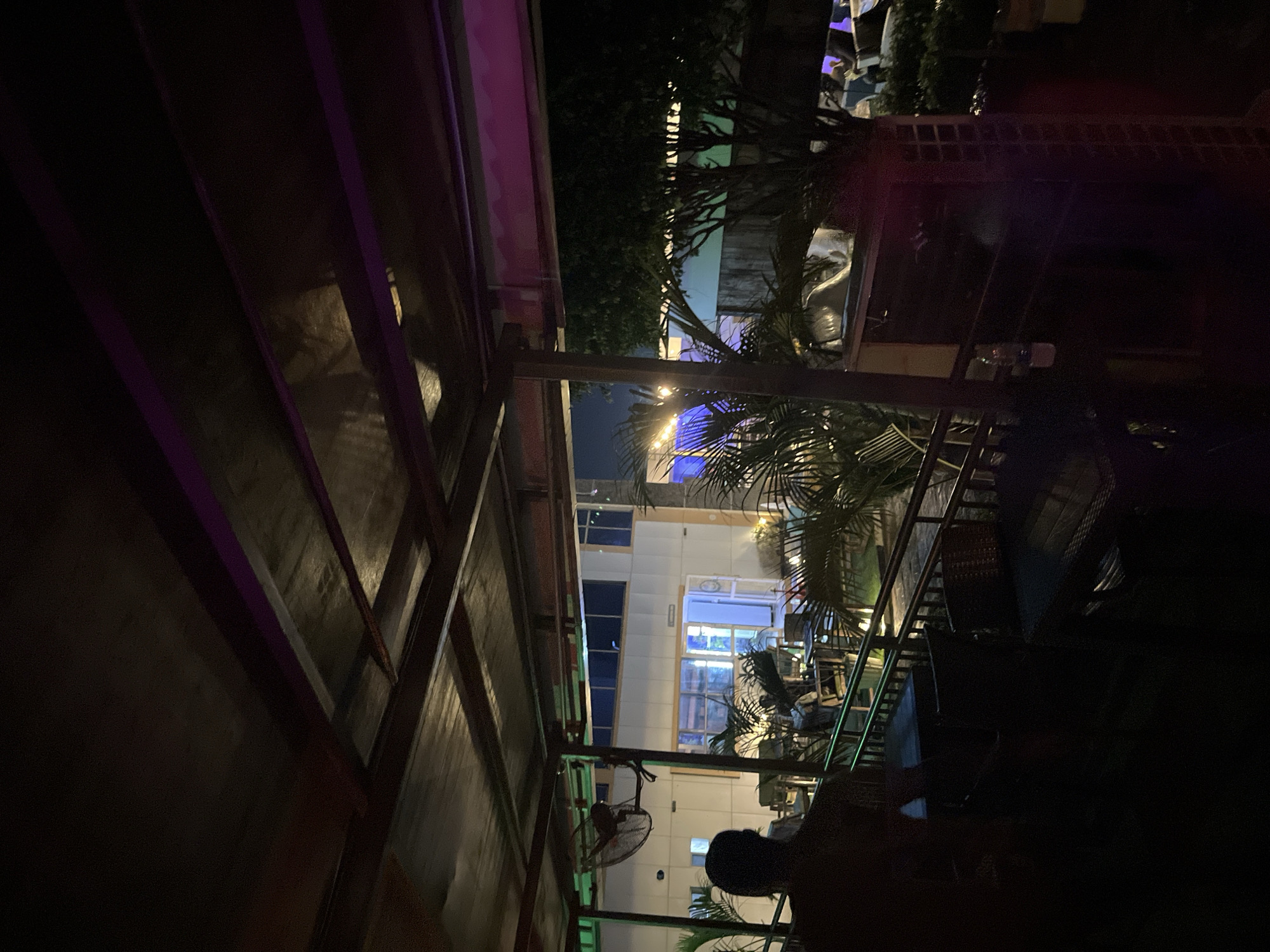 The image displays a nighttime scene within an outdoor or semi-enclosed dining/lounge area, located in Unknown, Unknown. The perspective is from under a dark, slatted roof structure, likely made of wood and corrugated metal, which occupies the upper portion of the frame. Purple LED strip lighting is visible along the underside of several roof beams on the right side.
In the immediate foreground, a person's back and head are visible in silhouette on the far left, seated at a dark, possibly wicker-style outdoor table. Additional dark, wicker-style tables and chairs are partially visible in the lower-left and center foreground. A metal railing extends horizontally across the mid-ground, separating the foreground seating from another patio section. A small, oscillating wall-mounted fan is attached to a support column on the left.
Beyond the railing, numerous potted palm-like plants are interspersed among more outdoor seating arrangements. This area is softly illuminated by warm white string lights visible on the left, and blue ambient lighting emanates from the background.
The background features a light-colored, multi-story building with multiple windows, some reflecting interior light. A prominent ground-floor entrance or window in the center of the building emits a bright blue glow. Dark tree silhouettes are visible against the night sky on the far right. The overall scene is dimly lit by various artificial light sources, creating a distinct evening ambiance.