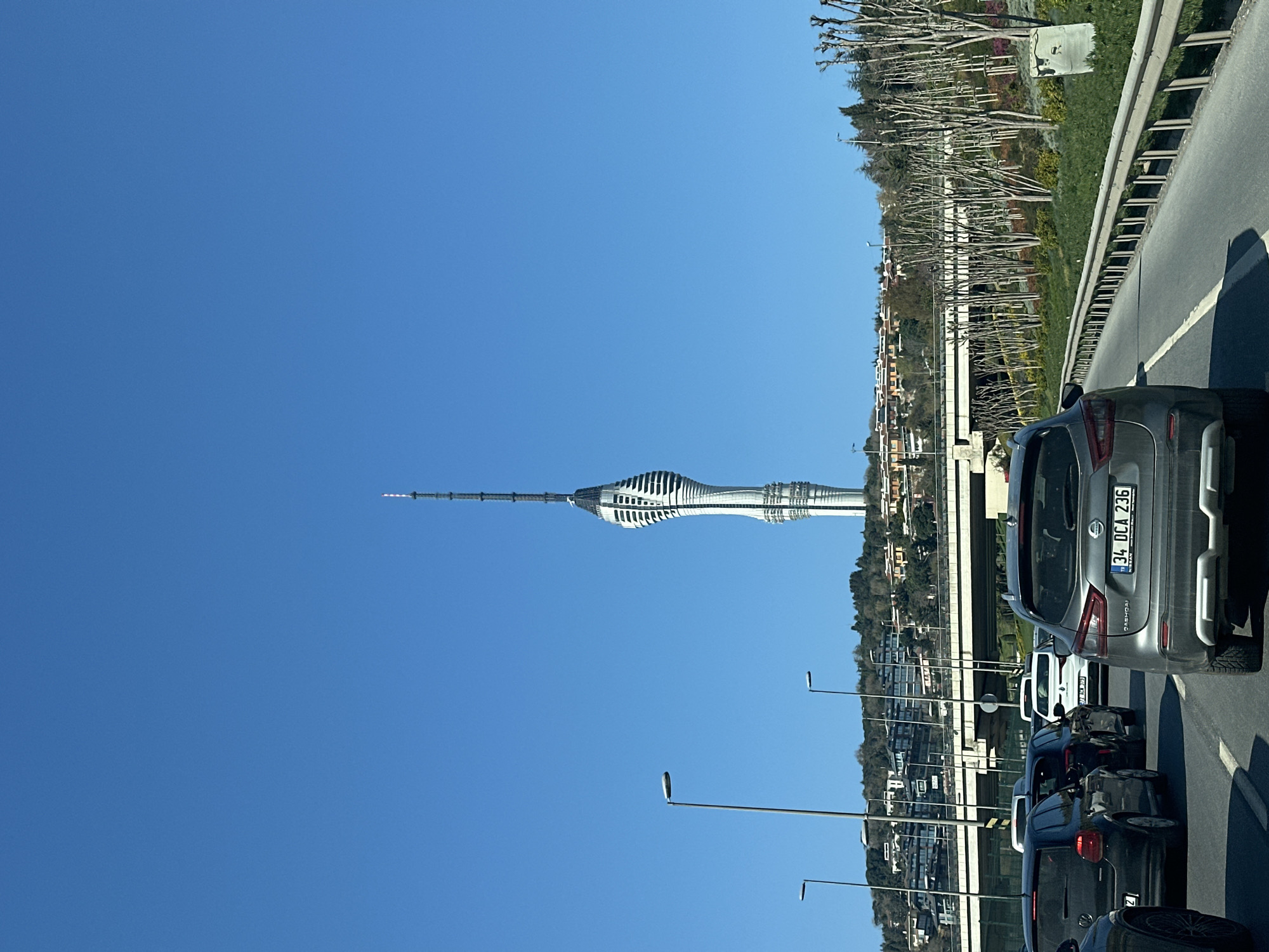 The image shows a traffic scene in Üsküdar, Turkey. In the background, the Çamlıca Tower is visible against a clear blue sky. The foreground shows several cars in traffic, with a silver Nissan Qashqai prominently displayed. The license plate of the Nissan reads "34 DCA 236". The cars are on a road with a barrier on the right side, beyond which is a grassy area with trees. Streetlights line the road. The image is rotated 90 degrees clockwise.