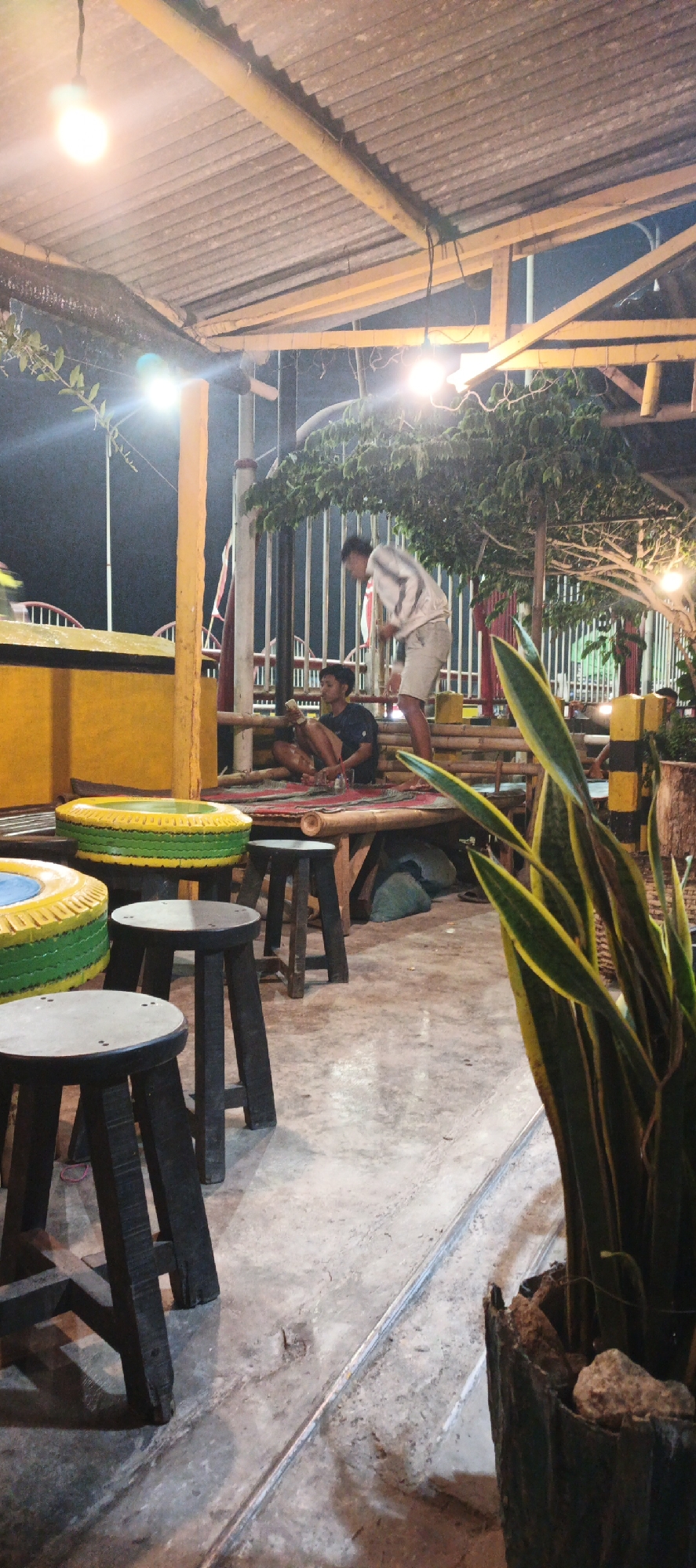 The image depicts an outdoor seating area at night. Two young men are visible, one sitting and the other standing and leaning over. The standing man is wearing a light-colored jacket and shorts, while the sitting man is in a dark t-shirt and shorts. They appear to be in conversation or engaged in an activity together.
The seating area is casual and appears to be outdoors, possibly a cafe or gathering spot. There are several low stools made of dark wood in the foreground. Behind the stools, there's a low bamboo table with a woven mat on top. Two tires, painted with green and yellow patterns, are stacked and serve as raised surfaces or tables. The ground is concrete.
In the background, there's a yellow structure, possibly a counter or wall, and a fence made of vertical wooden slats. Overhead, a corrugated roof is supported by wooden beams and illuminated by light bulbs. Lush green foliage, including a snake plant in a textured pot in the foreground and a tree with branches extending over the seating area, adds to the natural, relaxed atmosphere. The overall impression is a casual, possibly informal, outdoor entertainment venue. The image is taken in Kasiyan, Indonesia.