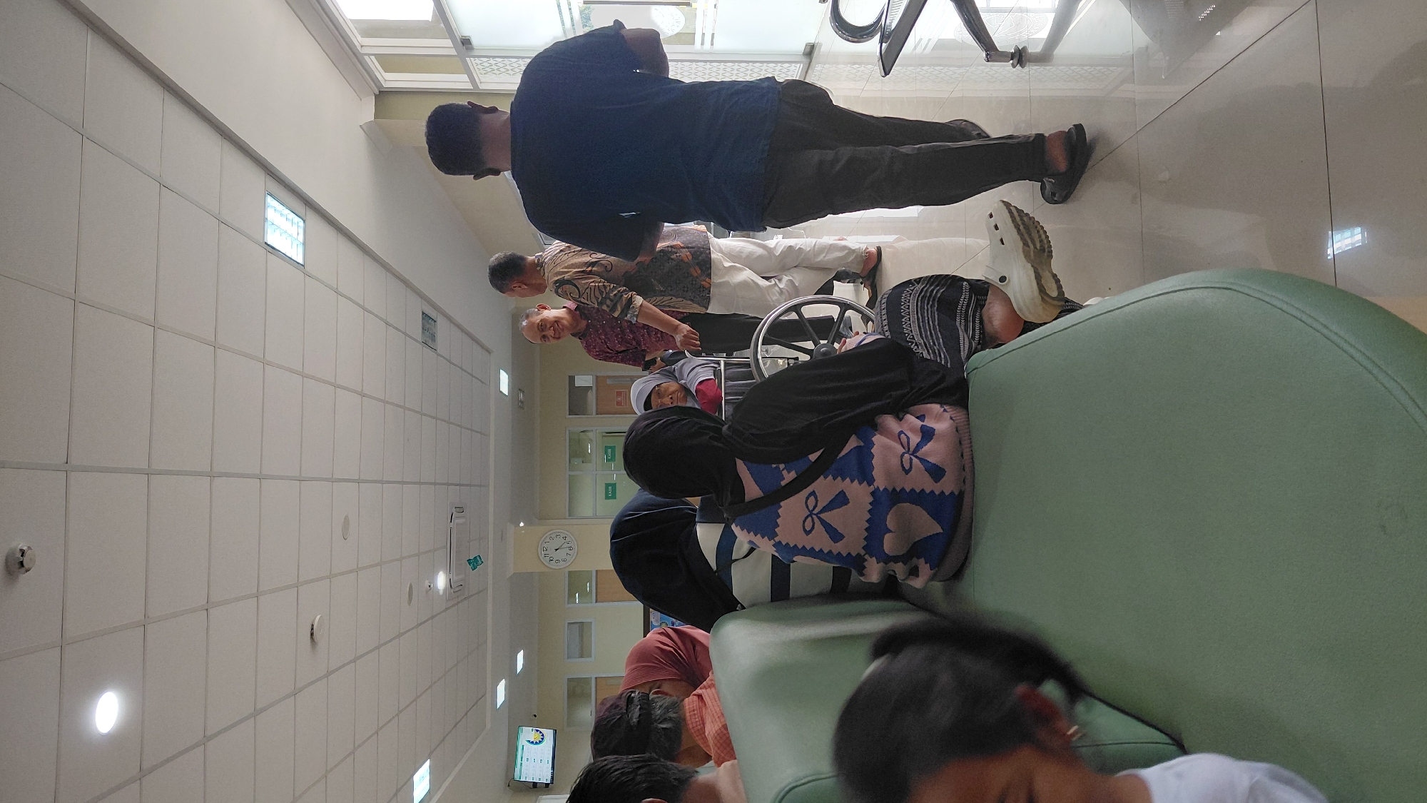 The image depicts an indoor waiting area, possibly a clinic or hospital, with several individuals present. In the foreground, a light green couch occupies the left side of the frame, with several people seated on it, their backs mostly to the camera. One person is wearing a blue and pink patterned sweater and a dark headscarf. To the right of the couch, a person in dark pants and a dark t-shirt walks away from the camera.
In the mid-ground, a group of three men are engaged in conversation. One man, wearing a dark patterned shirt, stands with his back to the camera. Another man, in a lighter patterned shirt and white pants, faces the first man and is smiling. A third man, in a maroon patterned shirt, is also facing the man in white pants and appears to be listening. Behind these men, a woman in a wheelchair is partially visible, wearing a light-colored headscarf and a red garment.
The environment appears to be a well-lit interior space with a tiled floor and a white drop ceiling with recessed lighting. A clock is visible on the wall, and there are signs that read "KASIR," which is Indonesian for "cashier." A monitor displaying a chart is mounted on the wall in the background. The overall scene suggests a public waiting area. The image is taken from a slightly elevated perspective, looking across the waiting area. Based on visual cues, such as the signage and the architectural style, this scene could be located in a public building in Rejasari, Indonesia, a district within the city of Denpasar, Bali.