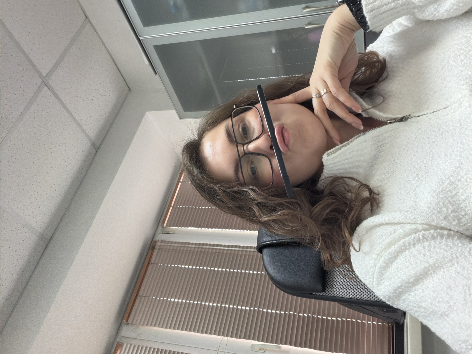An indoor portrait, likely a selfie, features a person, possibly a woman, with long, wavy brown hair, wearing dark-framed glasses. She is dressed in a white, textured, high-collared sweater with a visible zipper. A black, slender pen-like object is held horizontally between her nose and upper lip, suggesting a playful or contemplative pose. Her right hand is raised near her face, showcasing a ring on her ring finger and neatly manicured nails. A dark accessory is visible on her left wrist.
The person appears to be seated in a black office chair, part of which is visible at the bottom of the frame. The background indicates an office or commercial setting, with white acoustic ceiling tiles forming a grid pattern overhead. A light-colored wall or pillar is also visible. Further back, there's a window covered by horizontal blinds, suggesting daylight. Above and behind the person, dark, reflective cabinets or shelving units are visible. The overall impression is a casual moment captured in a professional or work environment. No text is visible in the image.