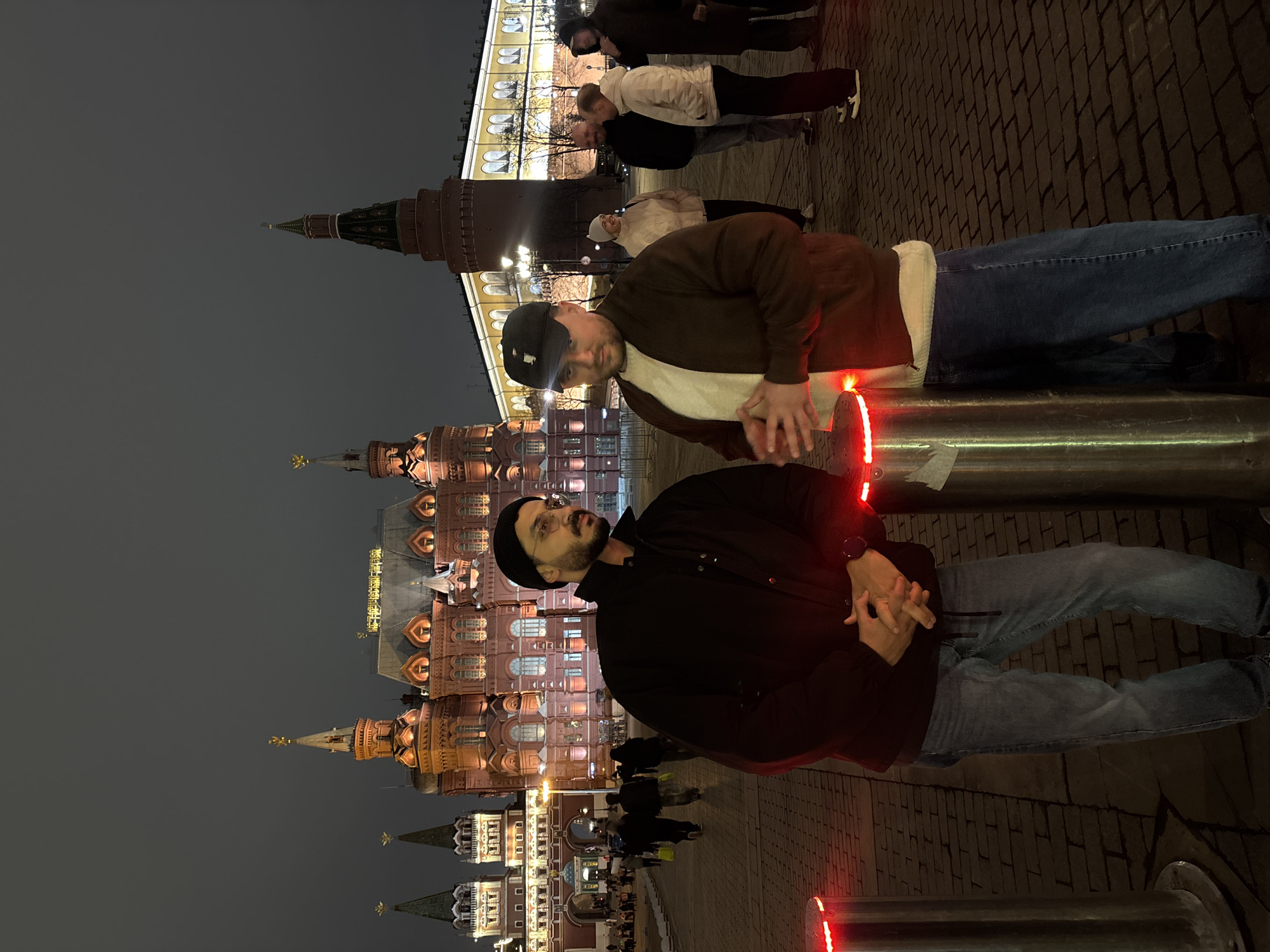 The image shows two men standing in a public square in Moscow, Russia, at night. In the background is the State Historical Museum, illuminated and prominently featured. The first man is wearing a black beanie, a black jacket, and jeans. The second man, positioned closer, is wearing a baseball cap, a brown jacket, and jeans. They are standing near metal bollards with red lights. People are walking in the background, and the sky is overcast. The ground is paved with brick.