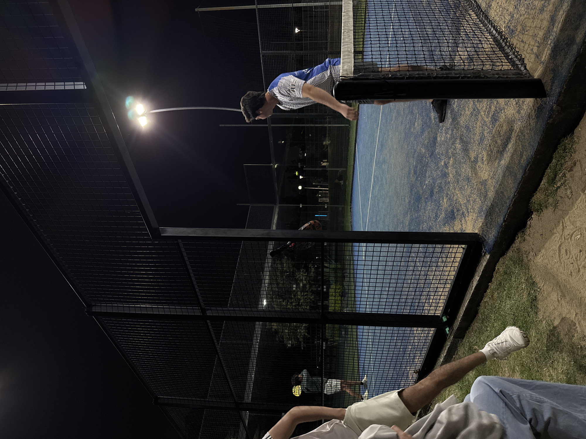 The image shows an outdoor padel tennis court at night in Heroica Puebla de Zaragoza, Mexico. The court has a blue surface and is surrounded by a black wire fence. A man in a white and blue shirt is leaning over the top of the fence, looking down at the court. At the bottom of the image, there are two people sitting on a bench, one wearing a white shoe. A streetlight illuminates the scene.