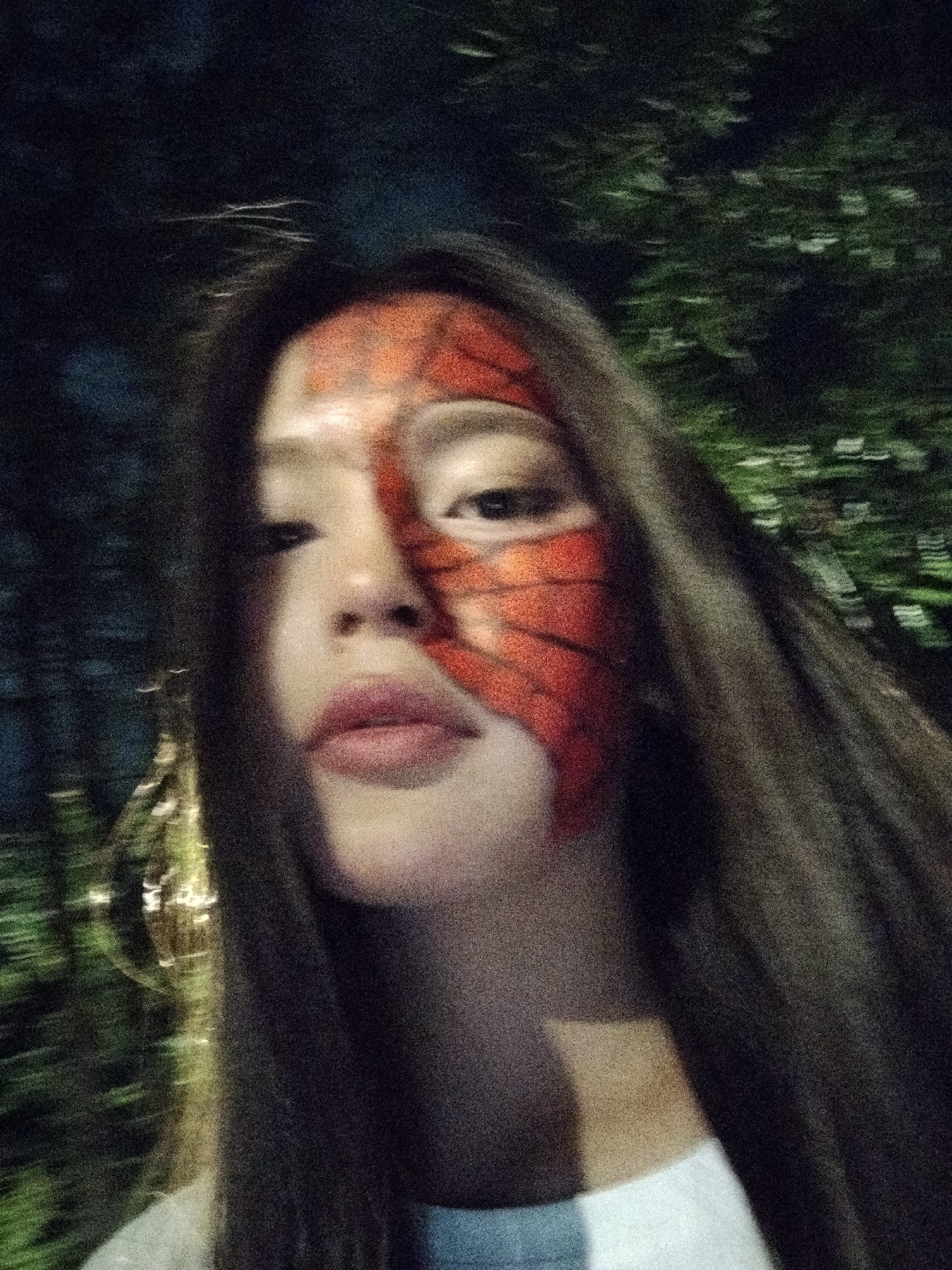 A young woman with dark hair and an orange and red spider web pattern painted on the right side of her face is captured in a close-up, blurry, nighttime photograph. She is wearing a white shirt. The background is a dark, out-of-focus depiction of trees and lights, suggesting an outdoor setting in Bishkek City, Kyrgyzstan.