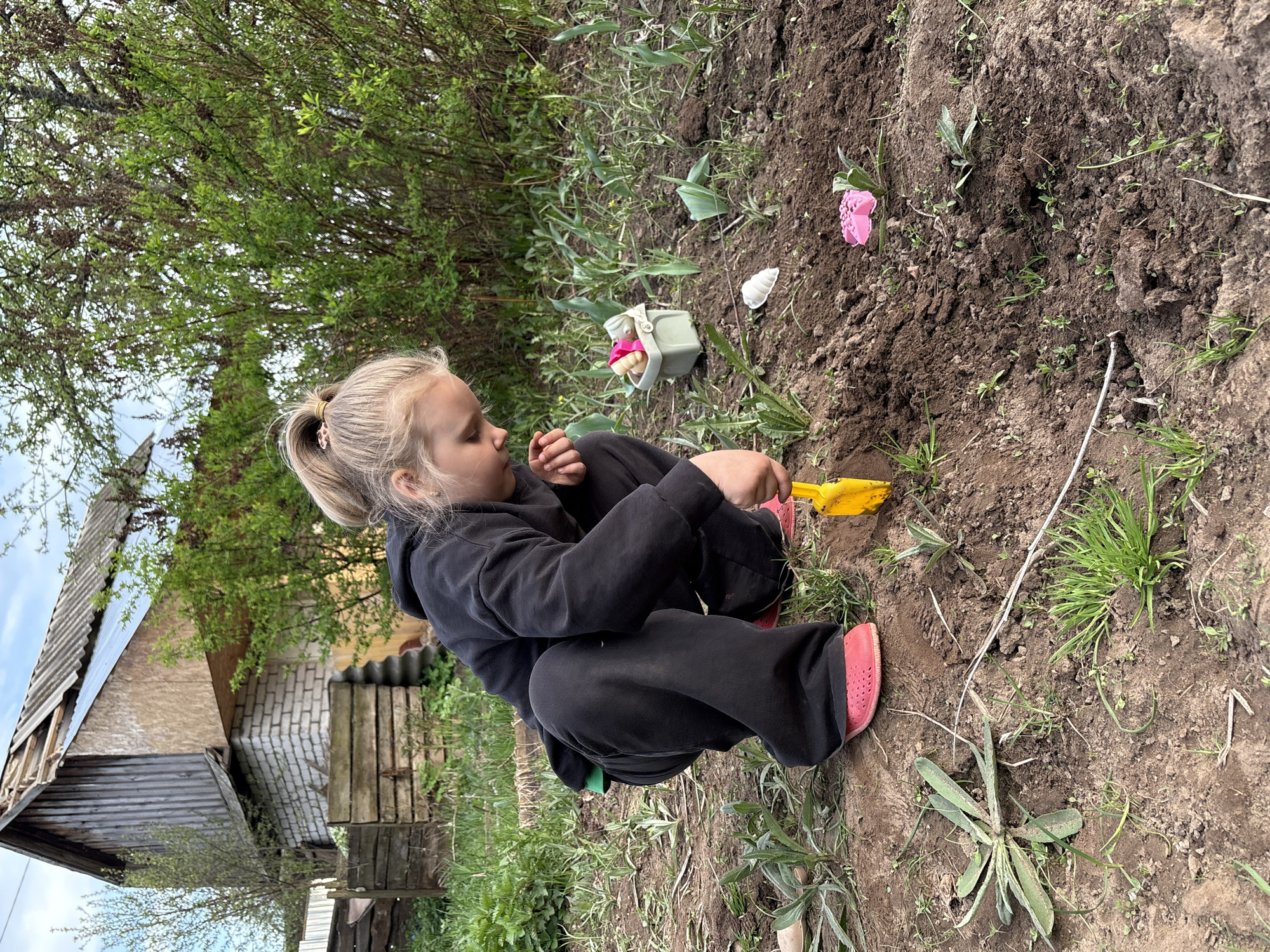 A young girl with blonde hair in a ponytail is playing in the dirt with a yellow toy shovel. She is wearing a black hooded sweatshirt, black pants, and pink Crocs. Beside her are a pink toy flower, a toy bucket, and a white seashell. The ground is dirt with scattered green plants and grass. Behind the girl is a bush and a partially dilapidated wooden building with a brick addition. The location is Chaikovsky, Russia.