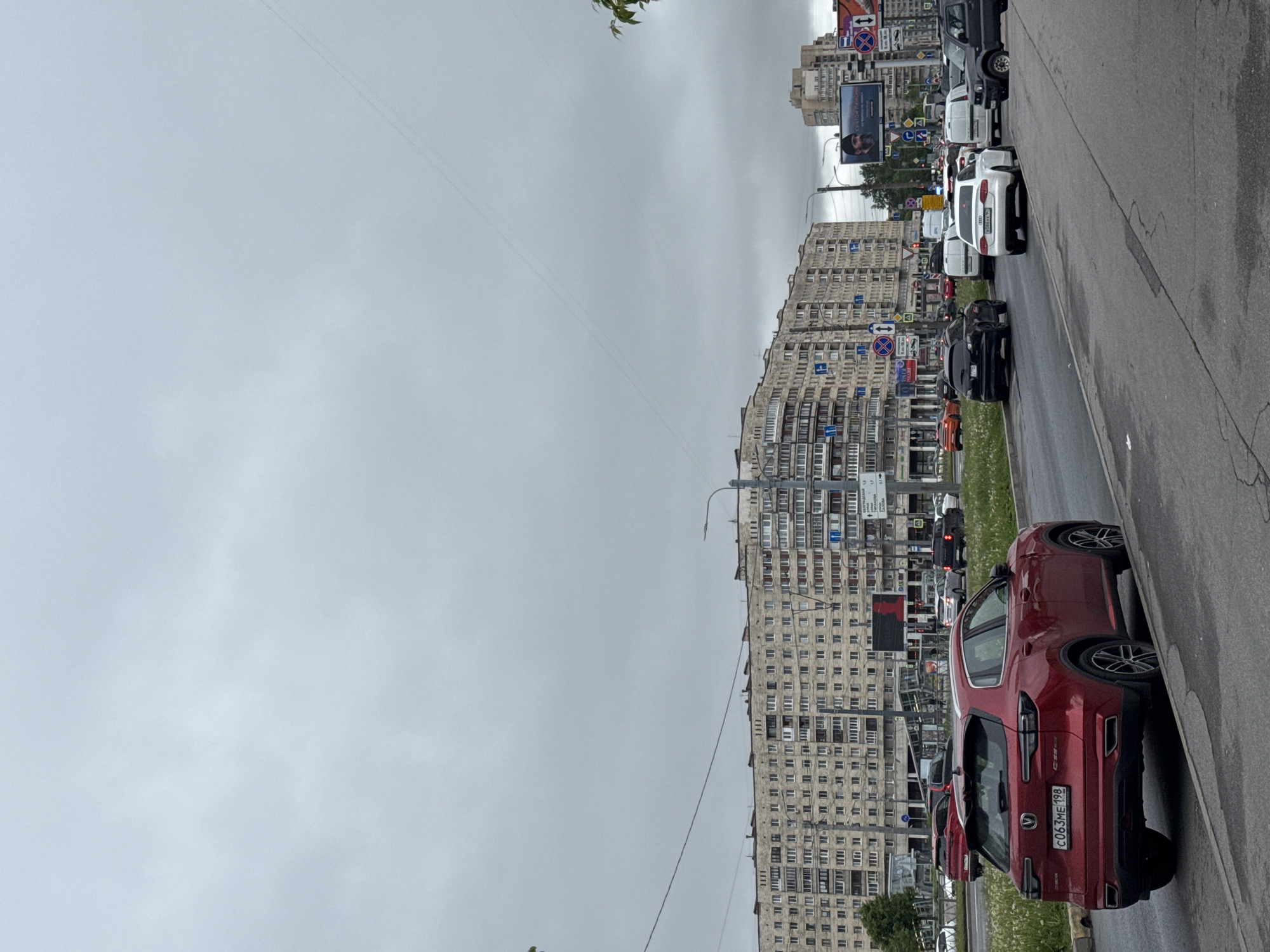 The image shows a street scene in Saint Petersburg, Russia, under an overcast sky. A large, high-rise apartment building with a repetitive pattern of windows dominates the background. The foreground features a red Changan CS75 parked on the side of the road, with its license plate number "C063ME 198" visible. A line of other cars, including white vans and a black SUV, are visible in the traffic lane. Street signs are present along the road, and a billboard is mounted on one of the buildings. The environment is urban, with typical city infrastructure like overhead wires and streetlights. There are no people explicitly visible in the frame.