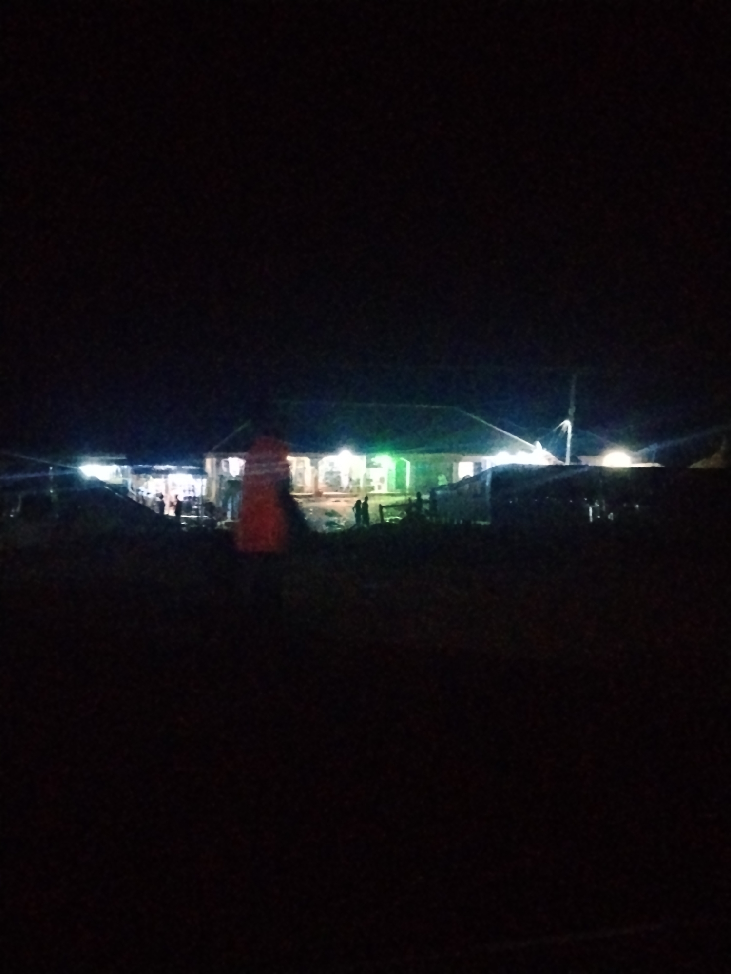 A nocturnal, low-light photograph captures a scene of activity in Durumi, Nigeria. In the mid-ground, a long, low-lying building is illuminated by bright artificial lights, appearing to be a marketplace or gathering space. Several figures, some of them indistinct due to the lighting and distance, are visible in and around the building. One prominent object in the foreground, to the left of the building, is a large, bright orange conical structure, possibly a traffic cone or a marker. Other structures, likely temporary stalls or shelters, are also present, and some are lit from within or by external lights. The foreground and background are largely dark, suggesting open space or unlit areas. The overall impression is of a busy night market or community event.