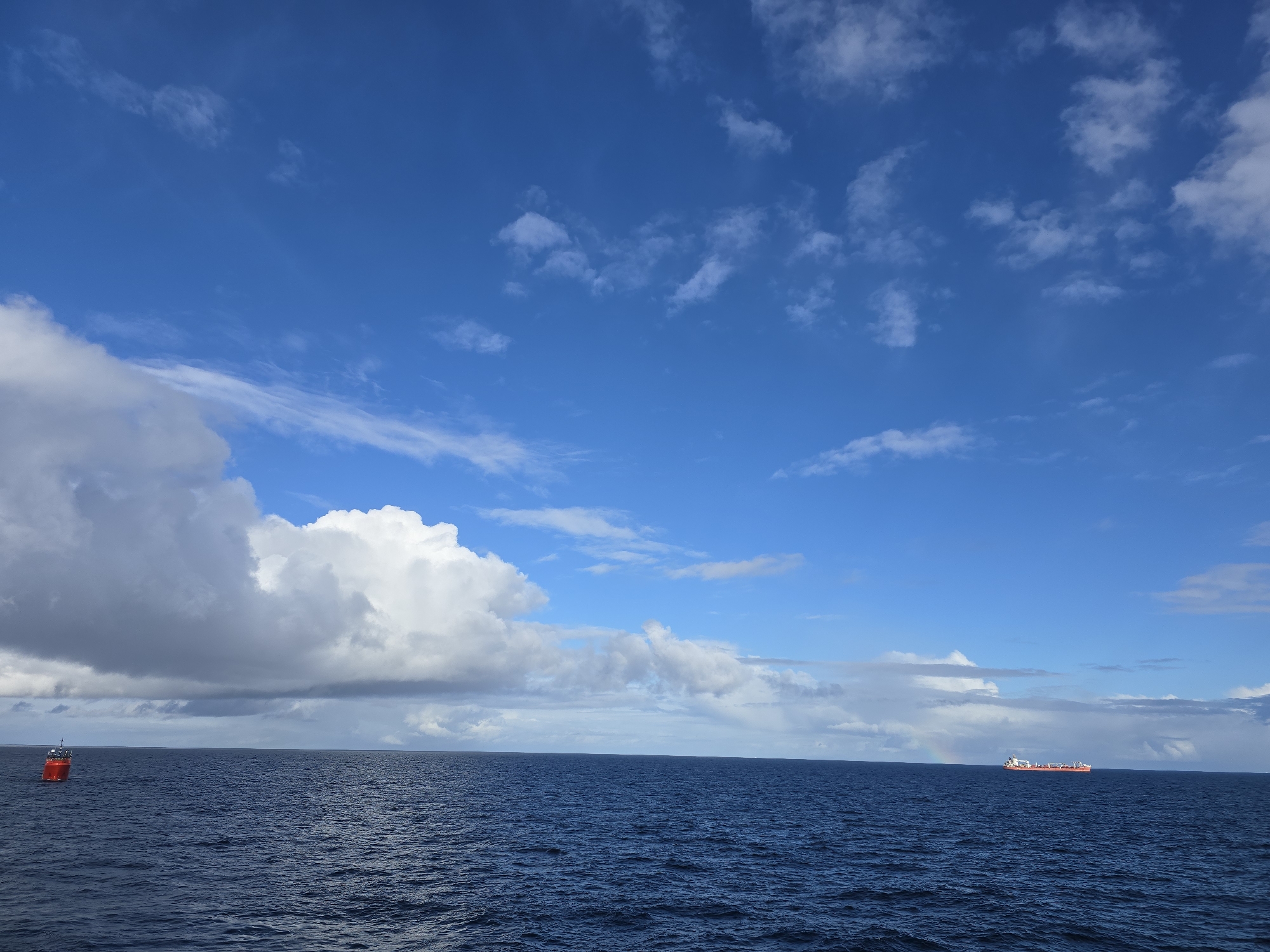 The image depicts a seascape under a partly cloudy sky. The sky transitions from a deep blue at the top to lighter shades near the horizon, dotted with scattered cumulus clouds. Dominating the left side of the frame is a large, puffy cloud formation, partially obscuring the horizon. The water is a deep blue, subtly textured by waves. On the left of the frame, a red buoy is visible in the water. In the distance, towards the right side, a ship is visible on the horizon. A faint rainbow arcs in the sky near the ship. The location is not identifiable based on the image content.