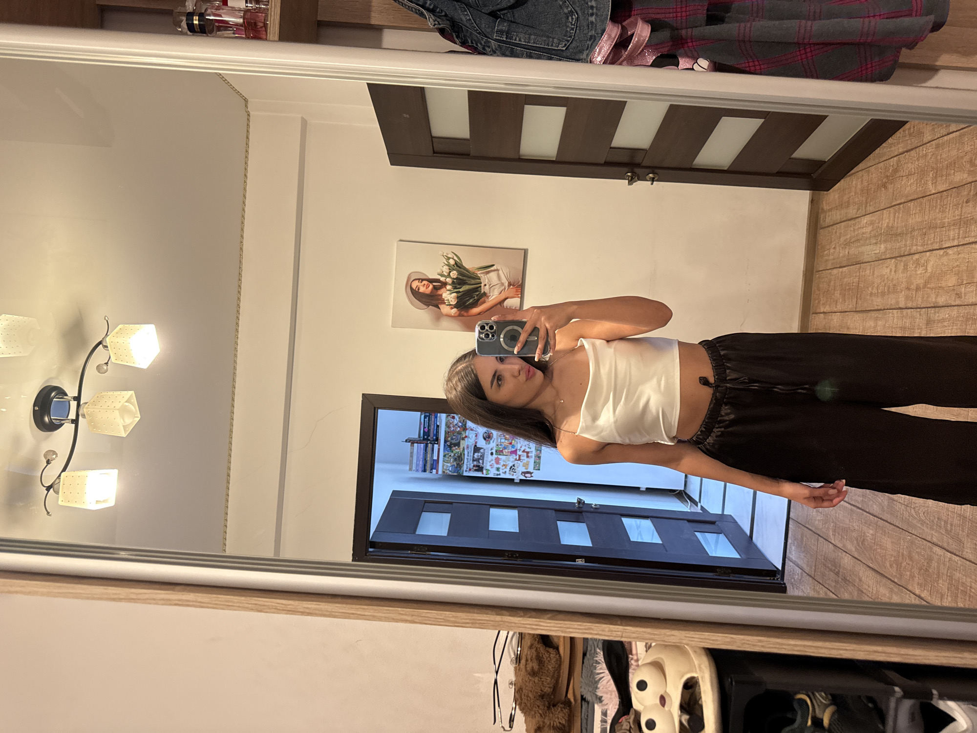 A young woman stands in front of a large mirror, taking a selfie. She is wearing a white satin crop top and black wide-leg pants. The mirror reflects a room with wooden floors and a dark wood door with frosted glass panels. To the left of the woman, in the mirror's reflection, a tall, narrow closet door is visible, and further left, a collection of shoes and a fluffy brown object are stacked on shelves. A framed picture of a woman holding tulips hangs on the wall to the right of the door. The room appears to be a bedroom or dressing area. The context of Chișinău, Moldova, is not discernible from the visual information provided in the image.