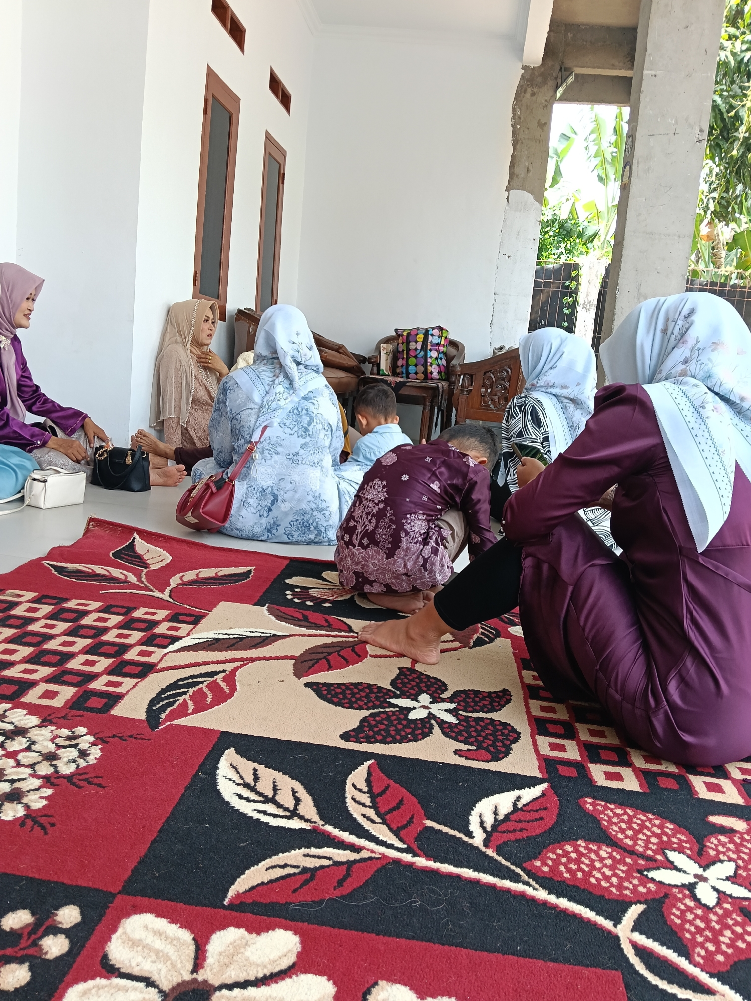 The image depicts a group of people, primarily women wearing hijabs, sitting on a patterned rug inside a building. The setting appears to be a home in Lemo, Indonesia, as indicated by the architectural style and surrounding foliage. Several women are seated, some engaged in conversation, while a young boy is seen crouching on the rug. The floor is covered with a large, brightly colored rug with floral and geometric patterns. In the background, there are wooden chairs and a view of outdoor vegetation through an open doorway. The lighting suggests it is daytime.