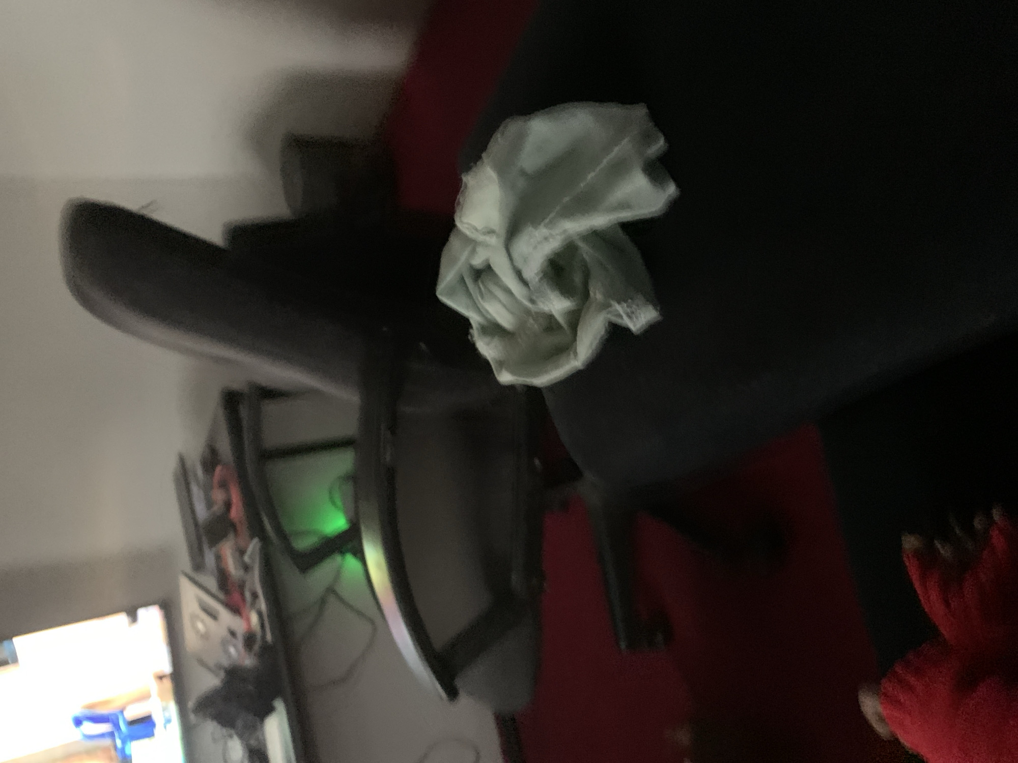 A close-up, dimly lit shot shows a cluttered desk with a computer monitor, a gaming chair, and what appears to be a folded piece of light blue fabric resting on a black surface. The fabric is crinkled and has some texture visible. The desk has various items on it, including what looks like a keyboard, a red object, and a dark object. Green light emanates from under the desk. The background is a plain wall. The lower part of the image reveals a person's feet wearing red socks, with their toes visible.