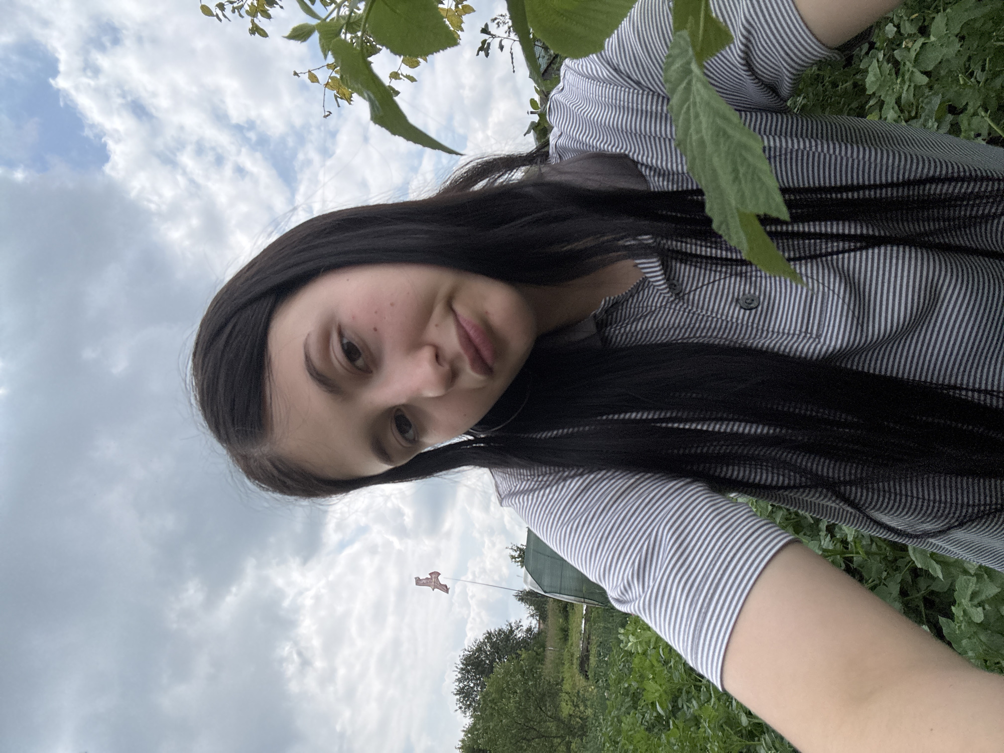 The image is a selfie taken outdoors in a rural or garden setting, likely in or near the city of Фоки, Russia, given the provided context. A young woman with long, dark hair and fair skin is centered in the frame, looking towards the camera. She is wearing a striped polo shirt. Her expression is neutral to slightly amused. The background consists of lush green foliage, suggesting a garden or field. In the distance, a greenhouse structure and a flag on a pole are visible. The sky is overcast with scattered clouds, indicating a daytime scene. The overall impression is that of a casual outdoor portrait.