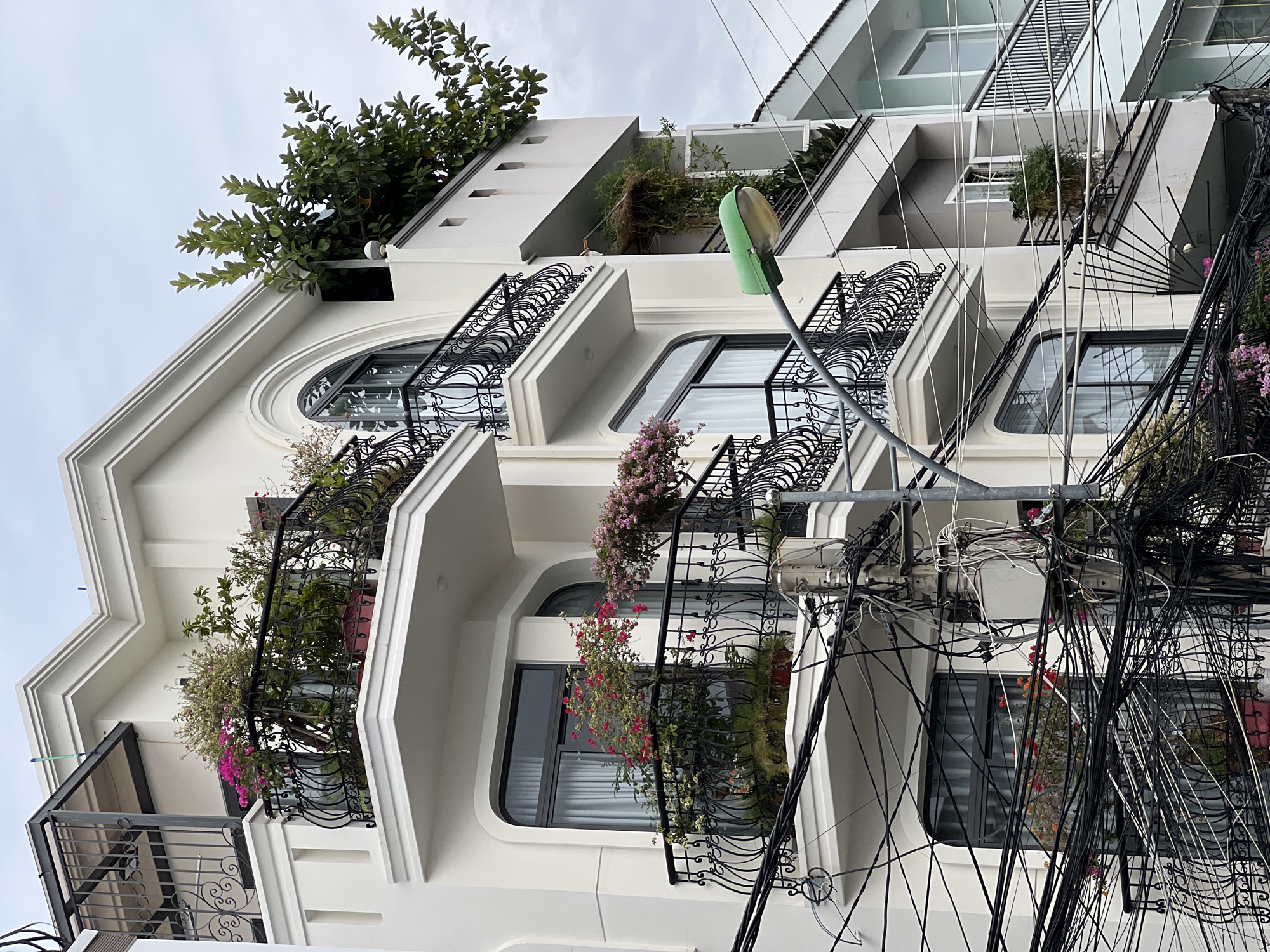 The image shows a white, multi-story building with ornate black balconies adorned with potted plants. The balconies feature decorative metalwork. On the upper floors, lush green trees are visible. The building is juxtaposed with a utility pole heavily laden with black electrical and communication wires, creating a dense, tangled network in the foreground. A green street lamp is also present. The architecture suggests a location in a tropical or subtropical urban environment. Based on the architectural style, greenery, and dense wire infrastructure, this scene is consistent with an urban setting in Nha Trang, Vietnam. There are no people visible in the image.
