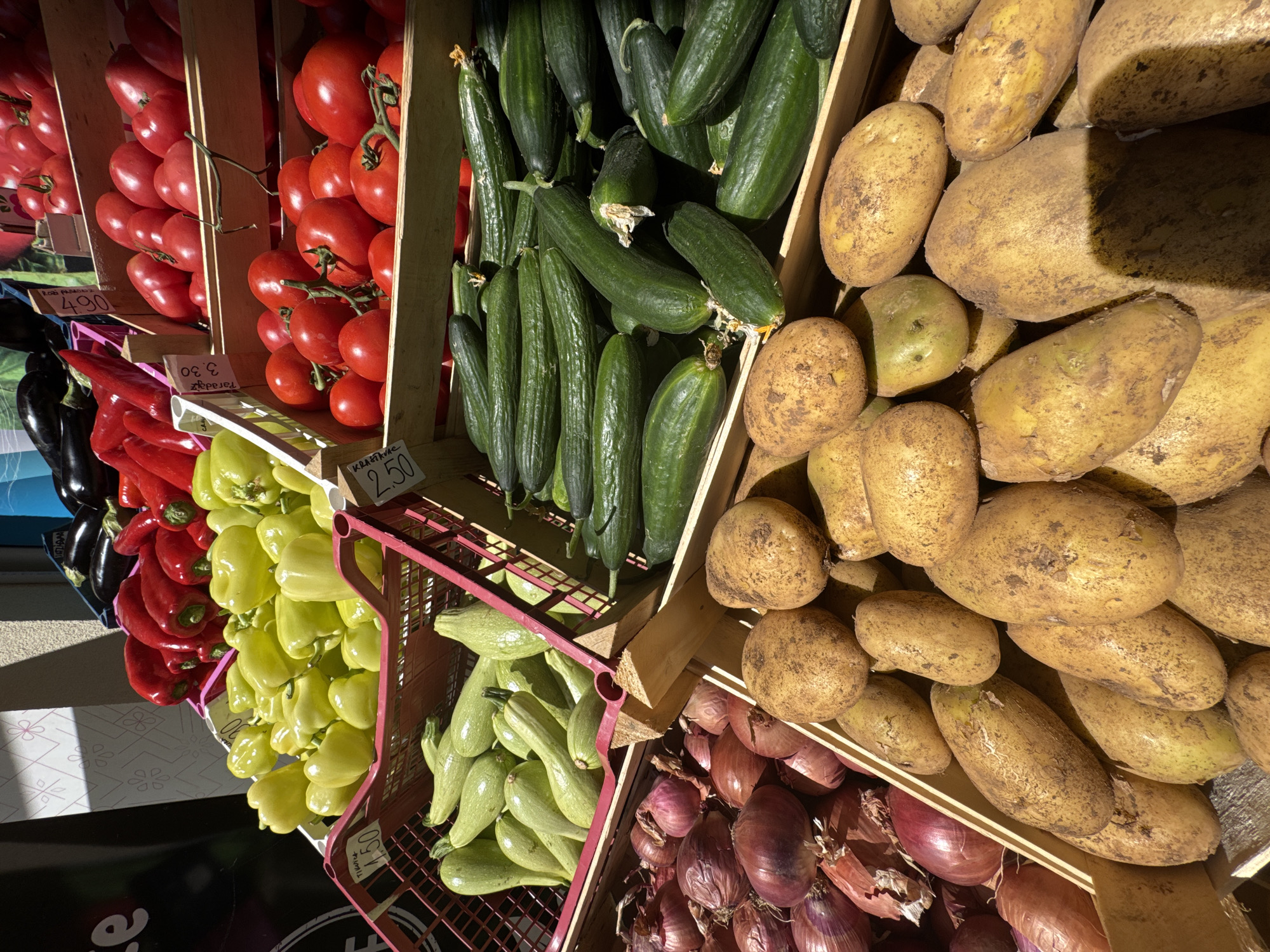 The image shows a display of various vegetables at a market stall in Bar, Montenegro. Tomatoes, cucumbers, potatoes, onions, zucchini, yellow bell peppers, red bell peppers, and eggplants are arranged in crates and baskets. Some price tags are visible with prices listed in local currency. The vegetables appear fresh. The scene is taken in daylight.