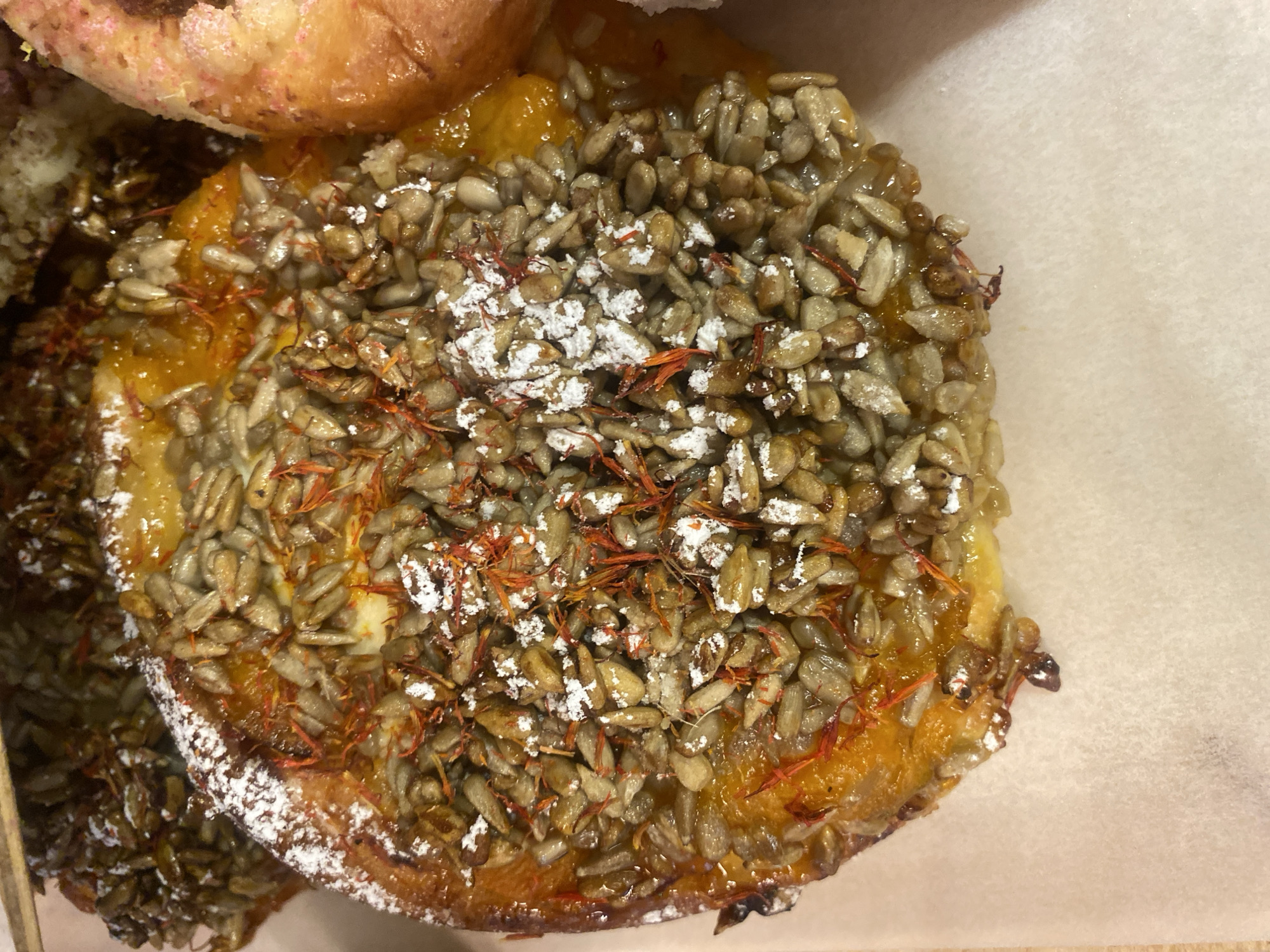 The media file shows a close-up, top-down view of a pastry on a light-colored, textured surface, likely parchment paper. The pastry is round and appears to be a tart or small cake. Its surface is covered with a dense layer of sunflower seeds, some of which are whole and some are broken. Interspersed among the seeds are fine, reddish-orange threads, possibly saffron or a similar spice, and a dusting of powdered sugar. The pastry itself has a golden-orange, glossy filling visible beneath the seeds, suggesting a fruit-based component like apricot or peach jam. The edges of the pastry show a baked crust. In the upper left of the frame, another pastry is partially visible, appearing lighter in color and possibly a different type, perhaps a brioche or croissant, with pinkish-white sugar crystals on its surface. The lighting is bright and appears to be from above, highlighting the textures of the seeds and the sheen of the filling. There is no visible text. The scene is an indoor, likely food-related setting, such as a bakery or cafe, and evokes a sense of freshly prepared or displayed baked goods. The focus is entirely on the food item, suggesting it is the primary subject of interest.
