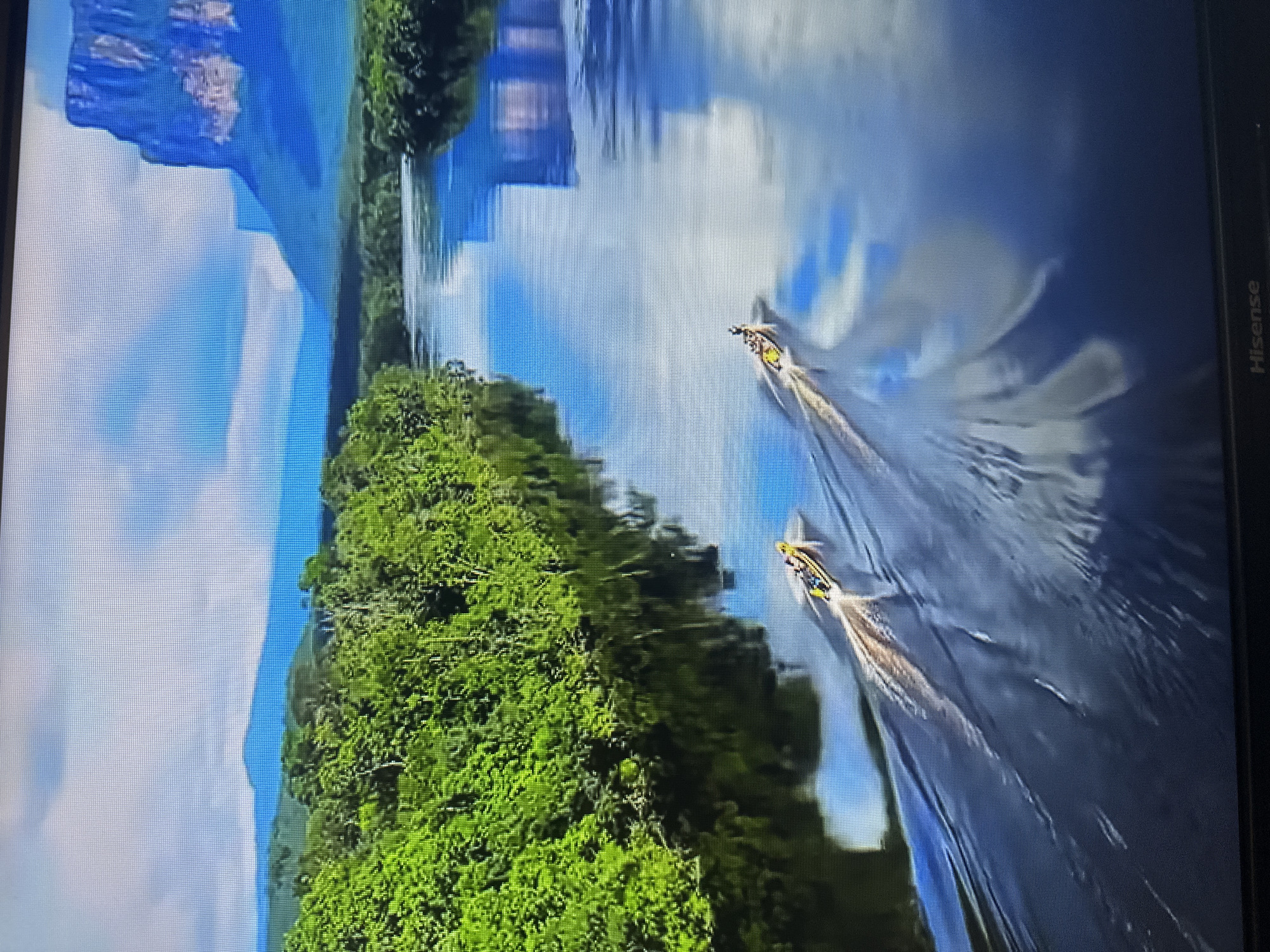 The image depicts a scenic landscape featuring a wide, calm river flowing through a lush green jungle. In the distance, towering, flat-topped mountains rise against a sky filled with dramatic clouds. Two yellow boats with people on board are seen cruising down the river, leaving wakes in their path. The water reflects the vibrant greenery of the jungle and the blue sky, creating a picturesque scene. There is no indication that this scene is located in Kyiv, Ukraine.