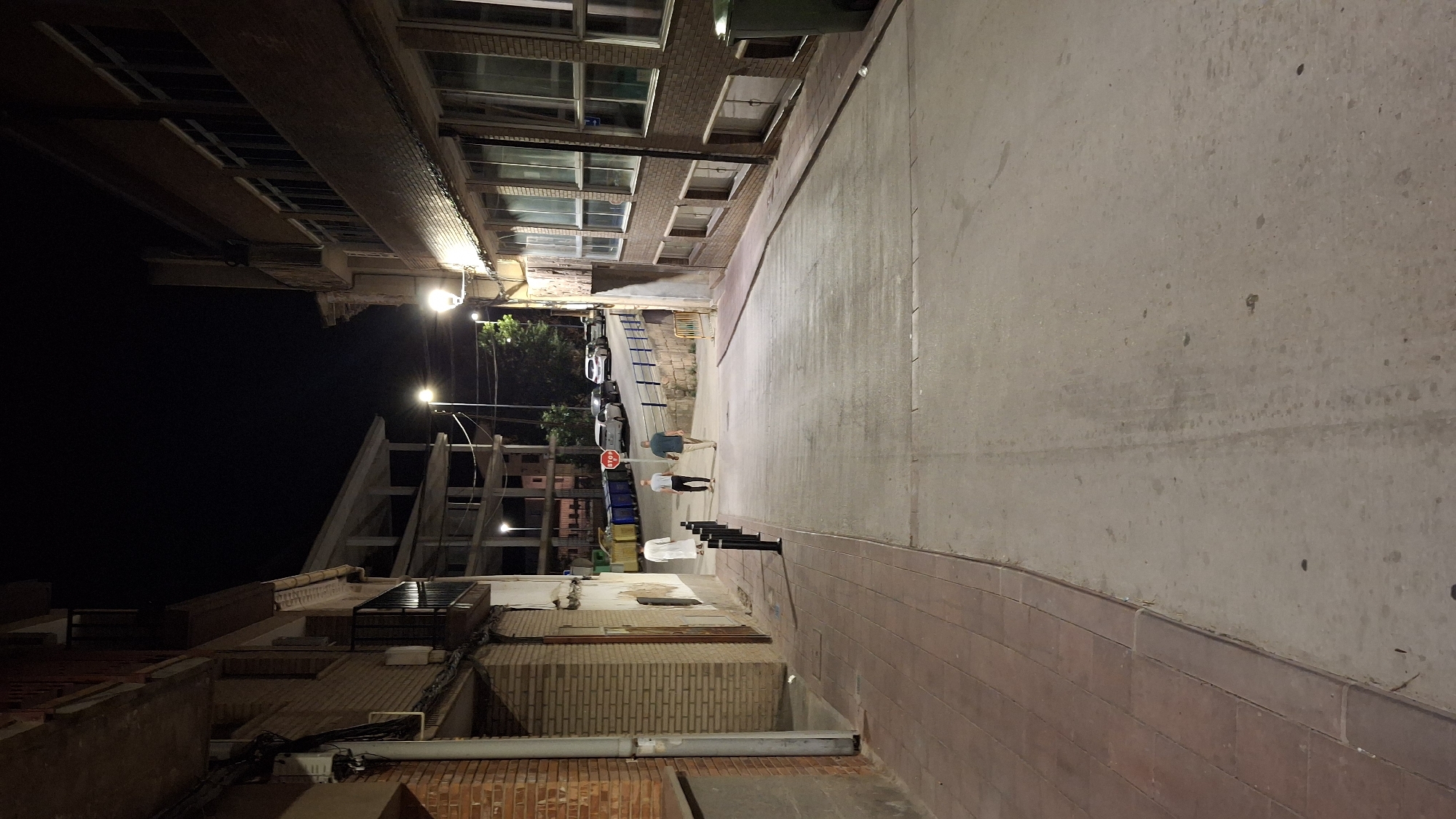 The image depicts a street scene in the town of Híjar, Spain at night. The perspective is from the middle of the street, looking uphill. On the left, a brick building with multiple balconies rises, featuring exposed brick and white plaster sections. A gray pipe runs vertically along its facade. The sidewalk on the left is paved with reddish-brown rectangular tiles, bordered by a darker curb.
On the right, another building with a brick facade and large, multi-paned windows is visible. Some of these windows have interior lighting. Further up the street on the right, a yellow barrier and a stone wall are present.
In the middle ground, three individuals are walking away from the camera, uphill. The person closest to the camera is wearing a long white robe. Behind them, two men are walking side-by-side; one is wearing a dark t-shirt and shorts, and the other is wearing a light blue t-shirt and dark shorts. Several cars are parked along the right side of the street further up, and a stop sign is visible near the parked cars. Streetlights illuminate the scene, casting shadows and highlighting the pavement, which is worn and shows tire marks in places. Several large blue and green trash bins are situated along the right side of the street. The sky above is dark, indicating nighttime.