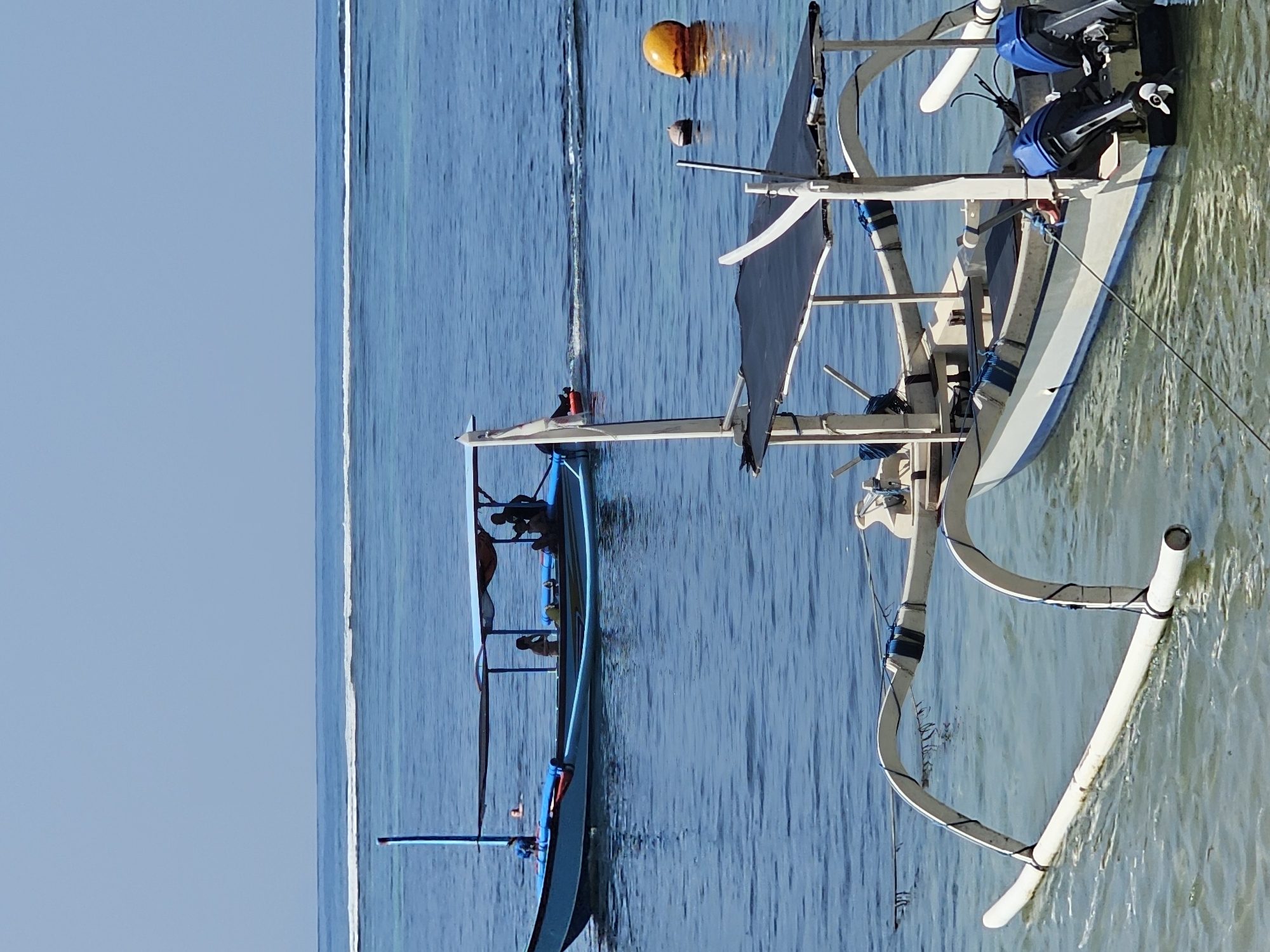 In Denpasar, Indonesia, on a clear day, two boats are visible in the calm, blue ocean near the shore. In the foreground, a white outrigger boat with a black canopy is moored. It features two blue outboard motors at the stern. In the background, a blue boat with a lighter blue canopy is moving away from the viewer. This boat also has people on board. The water is clear and shallow near the shore, with a distinct line of waves breaking further out. Two buoys, one orange and one brown, are floating in the water. The sky is clear and blue, indicating fair weather.