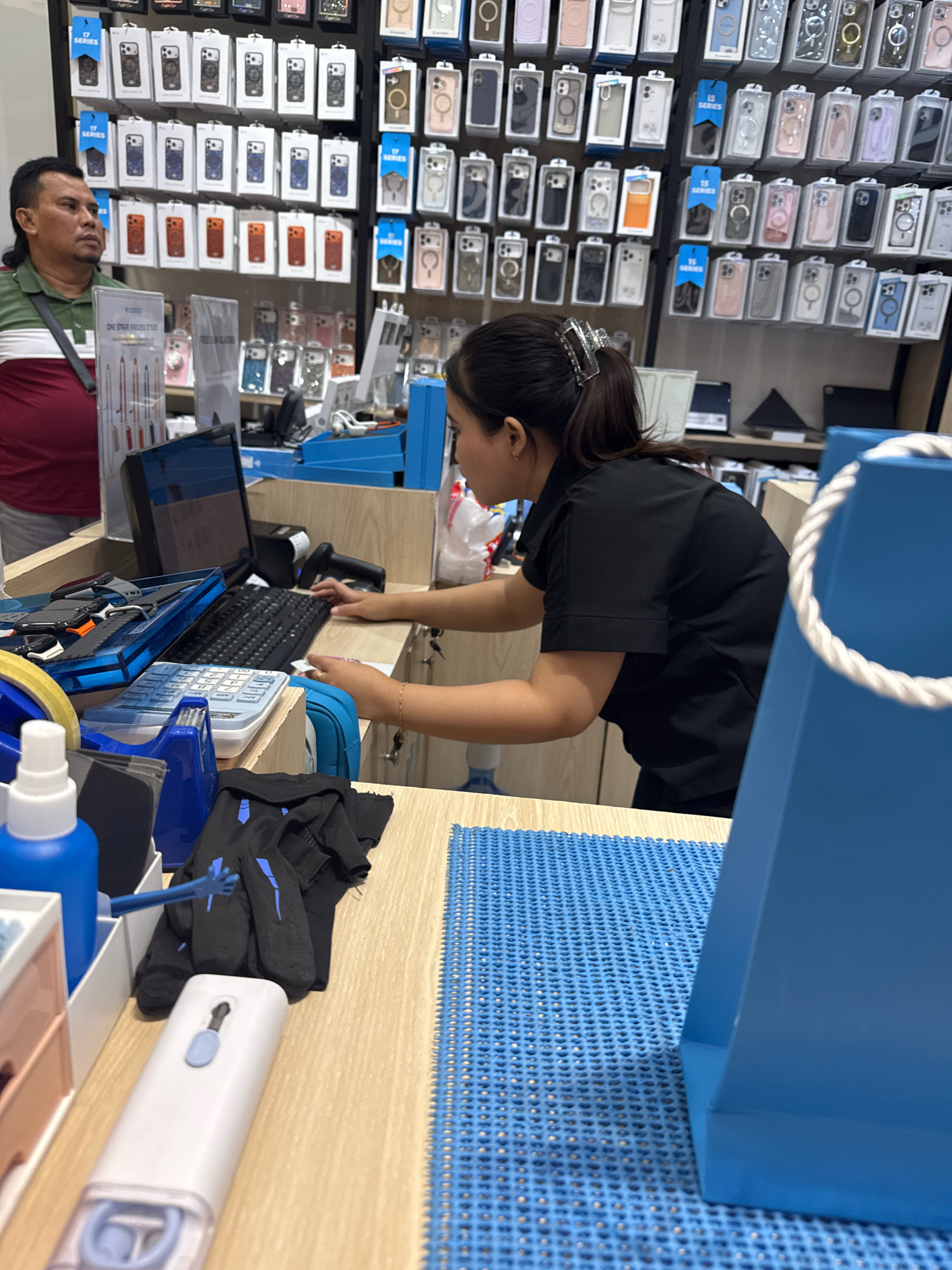 In a retail store in Surabaya, Indonesia, a male customer stands near a display of phone cases. A female employee is positioned behind a counter, operating a computer and keyboard, with a scanner and calculator nearby. The store appears to sell phone accessories and other electronic items, with shelves filled with various products. The environment is brightly lit and organized, typical of a commercial establishment.
