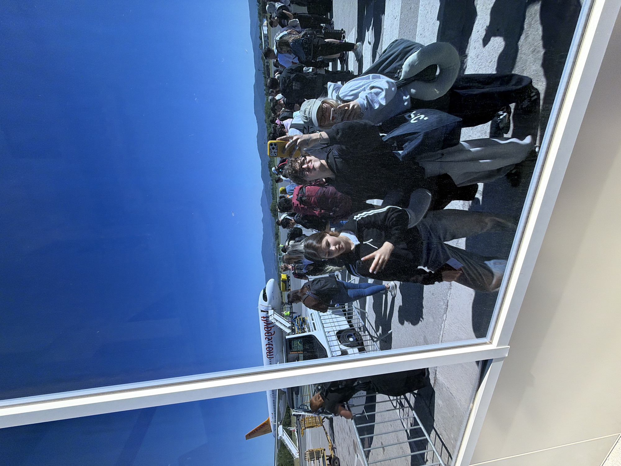 The image shows a photograph of a group of people boarding an aircraft. The photograph is hanging on a wall, and its reflective surface shows a second aircraft in the lower portion of the image. The people in the photograph are lined up next to the aircraft, presumably waiting to board. One person is taking a picture with a phone. The aircraft is a passenger plane with the words "flypgs.com" on the side. The photograph is of people boarding an aircraft at the Gornje Dubrave Airport in Bosnia and Herzegovina.