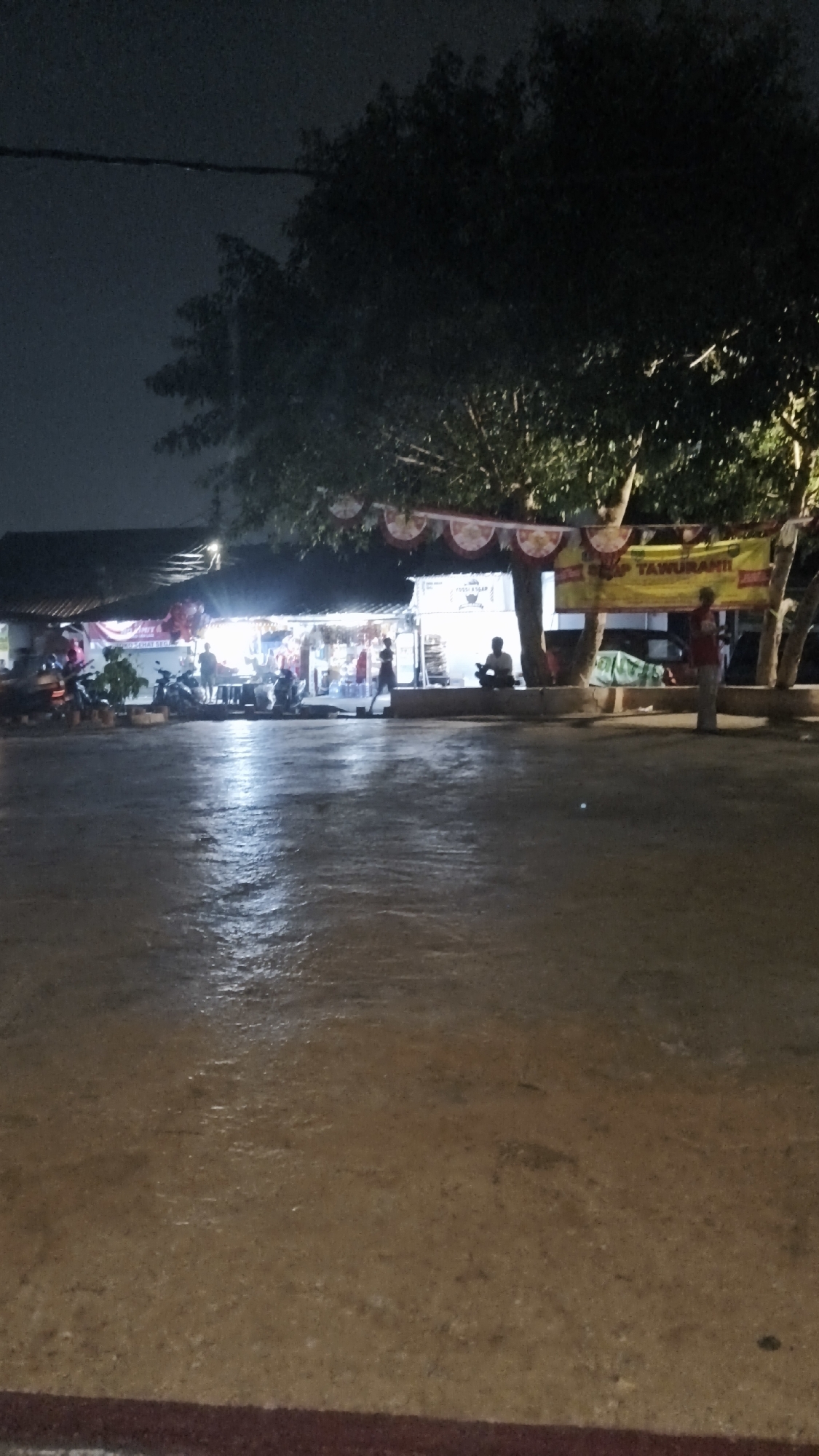 A vertical, low-angle, nighttime outdoor shot depicts a street-side market in Kebayoran Lama, South Jakarta, Indonesia. The foreground is a wet, reflective paved ground, likely from recent rain. In the midground, a row of stalls or small shops lines the left and center of the image. These establishments are illuminated from within, revealing goods displayed on shelves and tables. Several motorcycles are parked in front of the stalls. A few individuals are visible around the stalls, including one person standing in front of a stall on the left, another person walking in the center, and a person seated in front of a stall on the right. Overhead, festive red and white banners are strung between trees, adding a touch of decoration to the scene. A large yellow banner with text is also visible, hung from a tree in the right midground. The background is dominated by the dark silhouettes of trees against a dark, overcast sky. The overall atmosphere is that of a busy, albeit dimly lit, nocturnal marketplace.