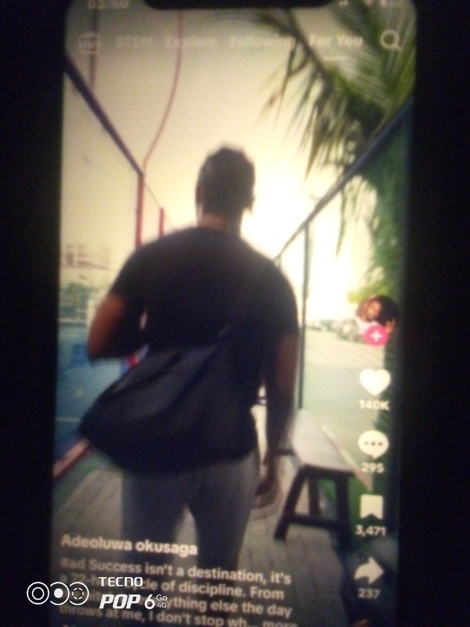 A vertical, slightly blurred, low-resolution photograph captures a person from behind, walking away from the camera on a pathway. The person is wearing a dark t-shirt and light-colored pants, with a dark messenger bag slung over their right shoulder. The pathway appears to be outdoors, possibly a court or recreational area, with a railing and some palm fronds visible in the background. The setting is bright, suggesting daylight.
On the right side of the image, a social media interface is overlaid. This includes icons for likes, comments, shares, and possibly other engagement metrics. Numbers such as "140K", "295", "3,471", and "237" are visible next to these icons, indicating engagement on a post. Text overlays also appear, including a username or caption with the name "Adeoluwa okusaga" and the phrase "Success isn't a desti...". Below this, a logo with the text "TECNO POP 6" is visible.
The specific location within Howayi, Nigeria cannot be determined from the visual information provided. The image depicts a digital interface on a mobile device, not a direct view of a specific geographical location in Howayi.