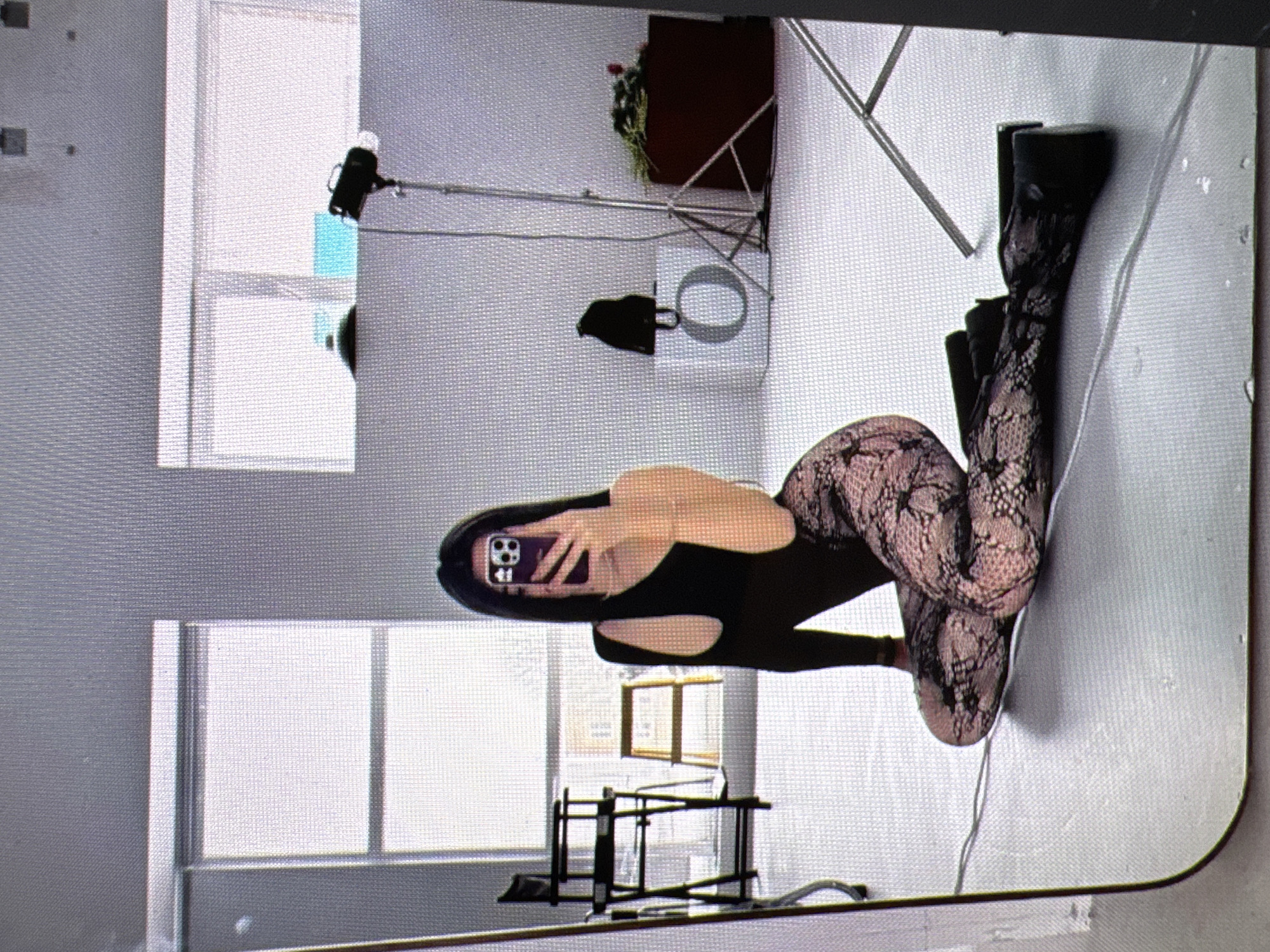 A young woman with long dark hair is posing for a photo in a studio. She is wearing a black one-shoulder top and patterned tights. The studio has large windows, studio lights, a makeup chair, and some props. The woman is holding a phone up to take a selfie. The scene is likely indoors, possibly in a professional photography studio. The woman is sitting on the floor with her legs crossed. Her body is angled towards the camera and she is looking at her phone screen.